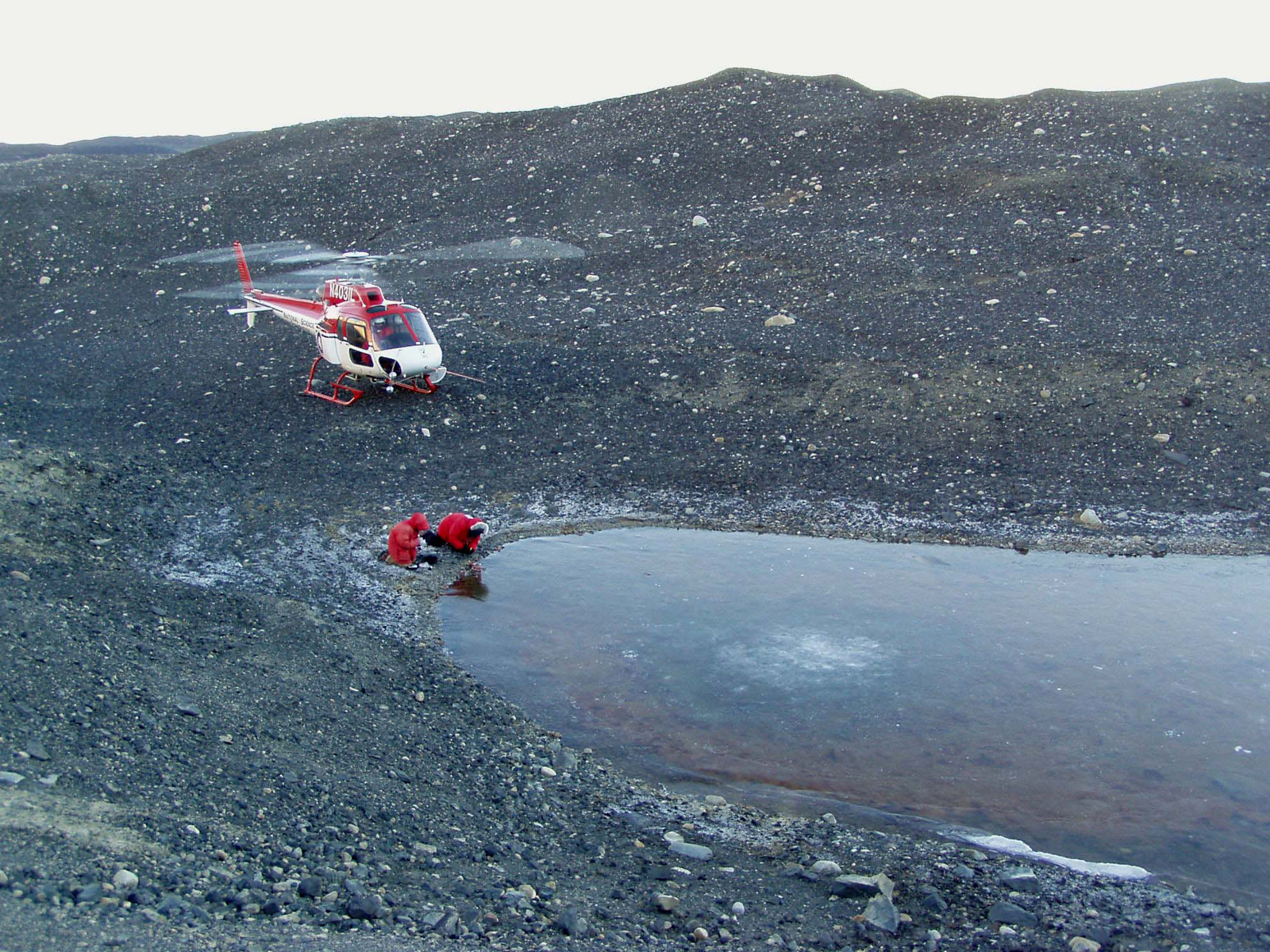Two people kneel near a small lake while a helicopter waits nearby.