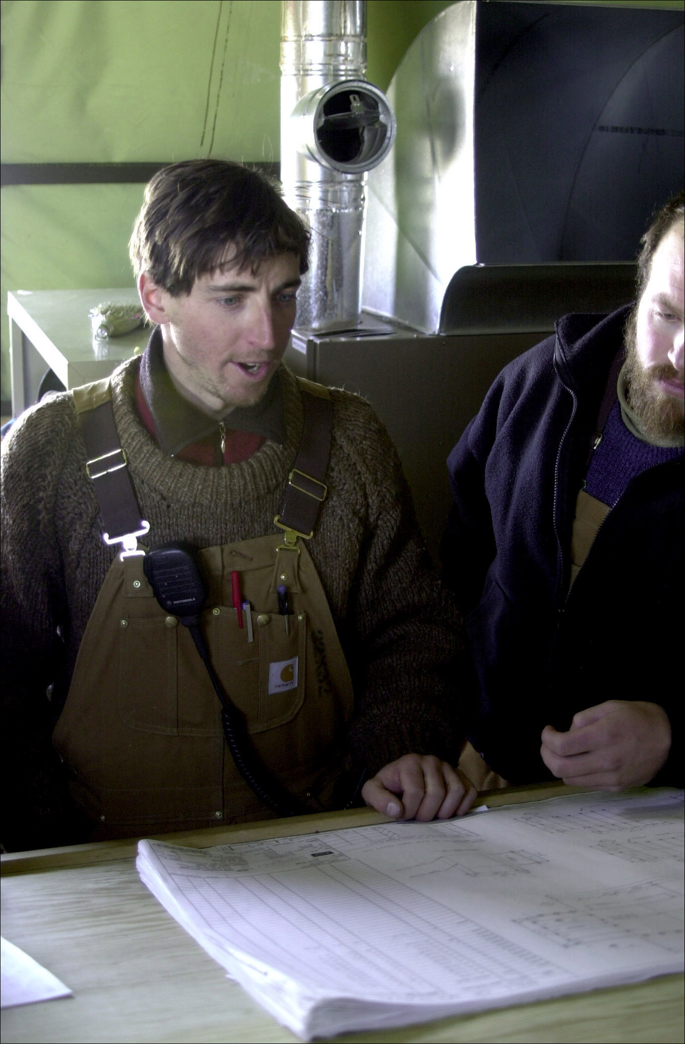 Two men sit at a table inside a hut, discussing the construction plans on the table.