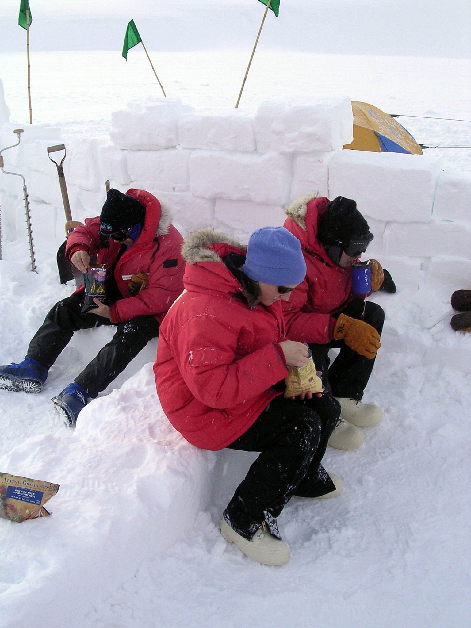 Three people in red parkas in front of a wall made of snow.