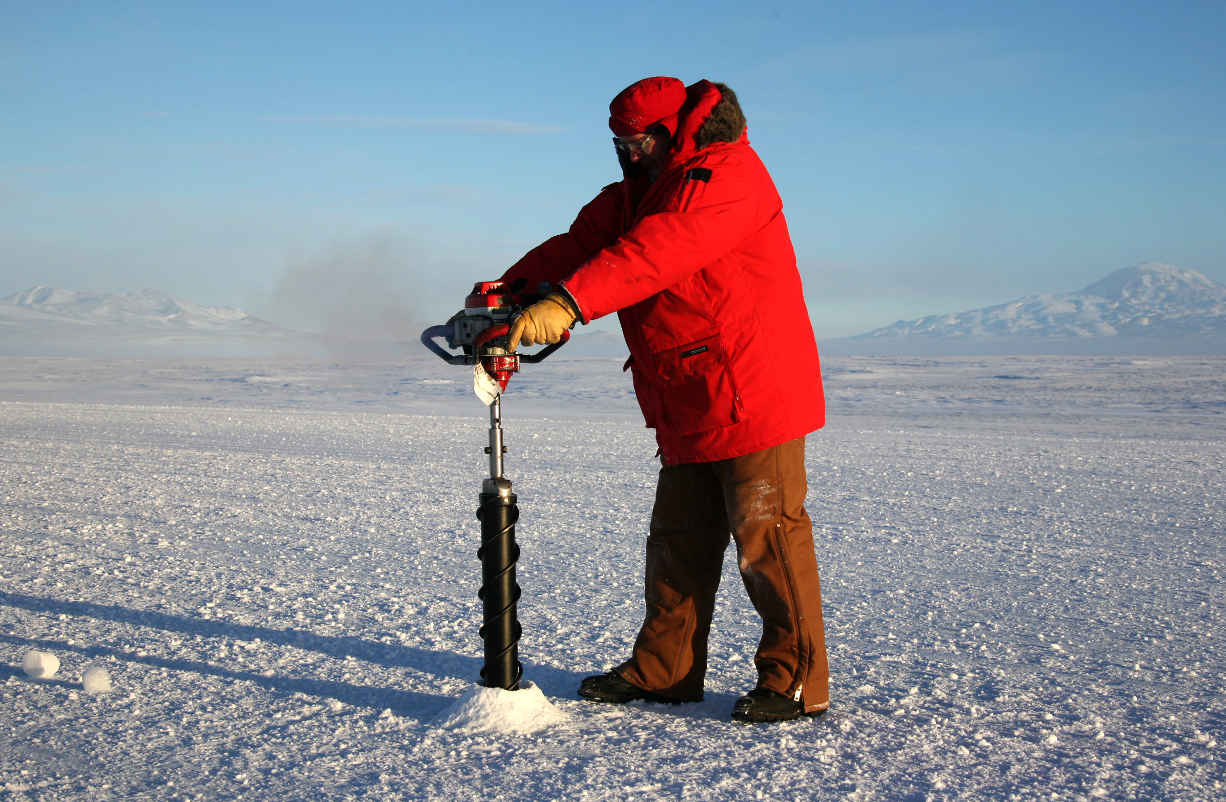Man in red coat drilling into ice.
