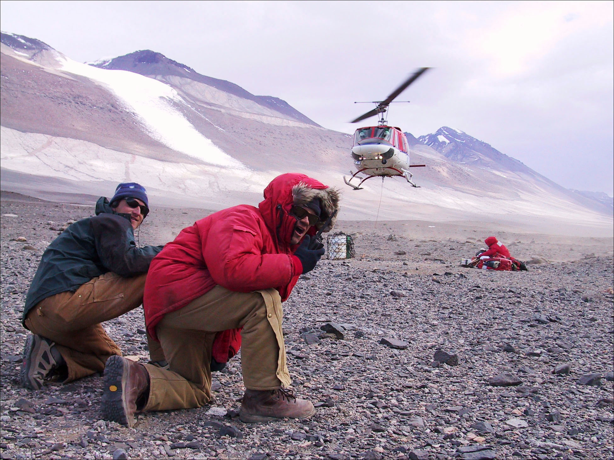 Two men crouch to the ground as a helicopter lands nearby.