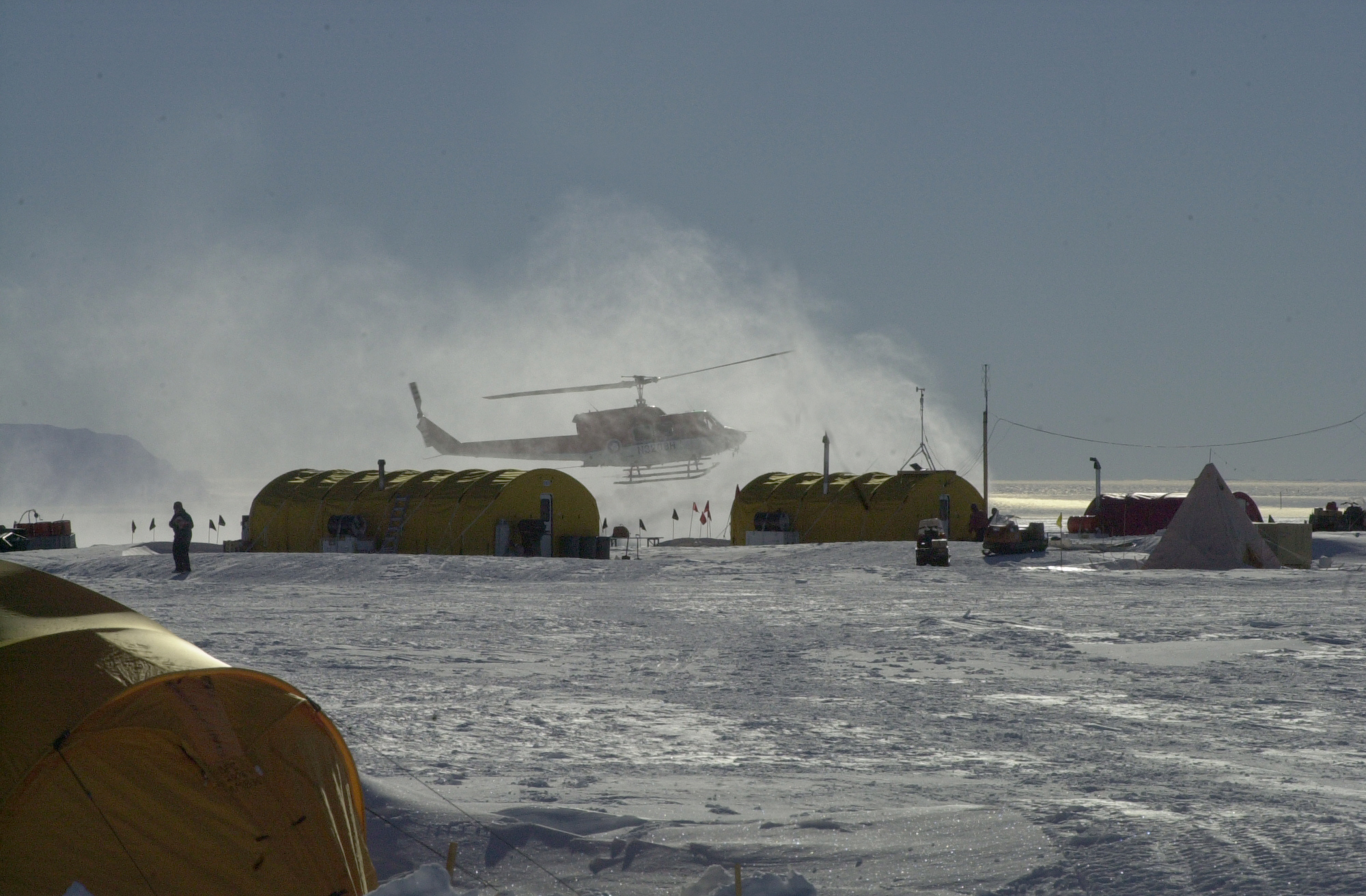 A helicopter landing on a snowy field near temporary buildings.
