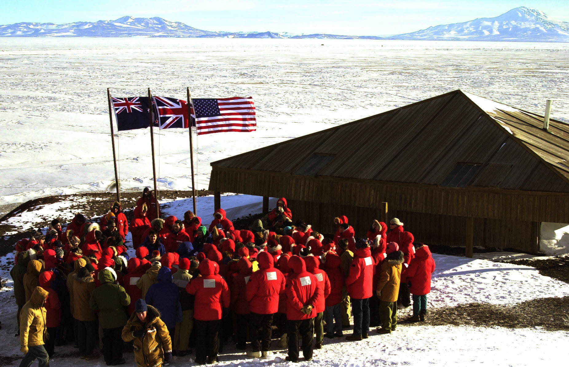 About 30 people in red parkas stand near an old hut during a ceremony.