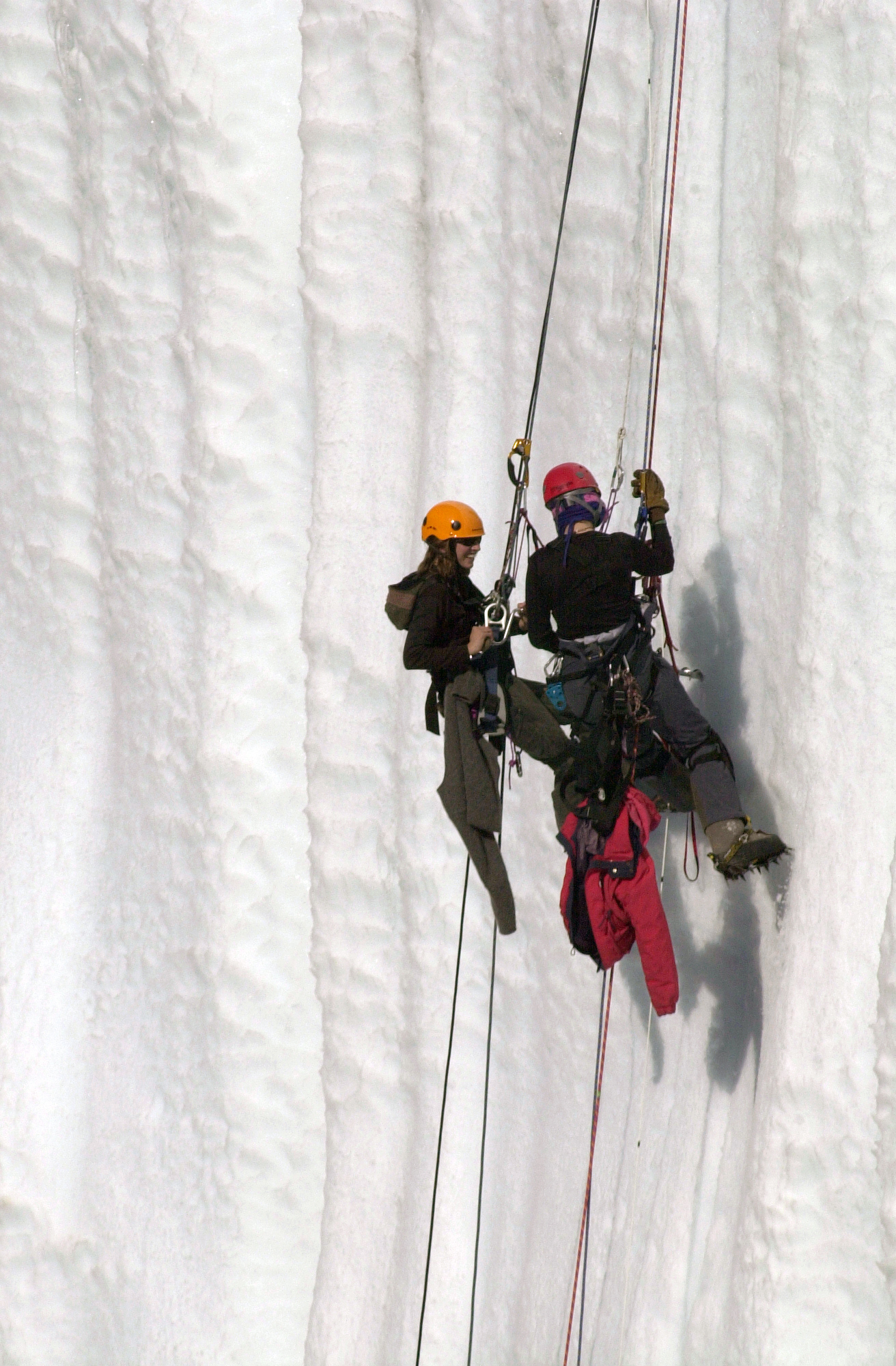 Two people in full climbing gear hang from ropes on a snow wall. 