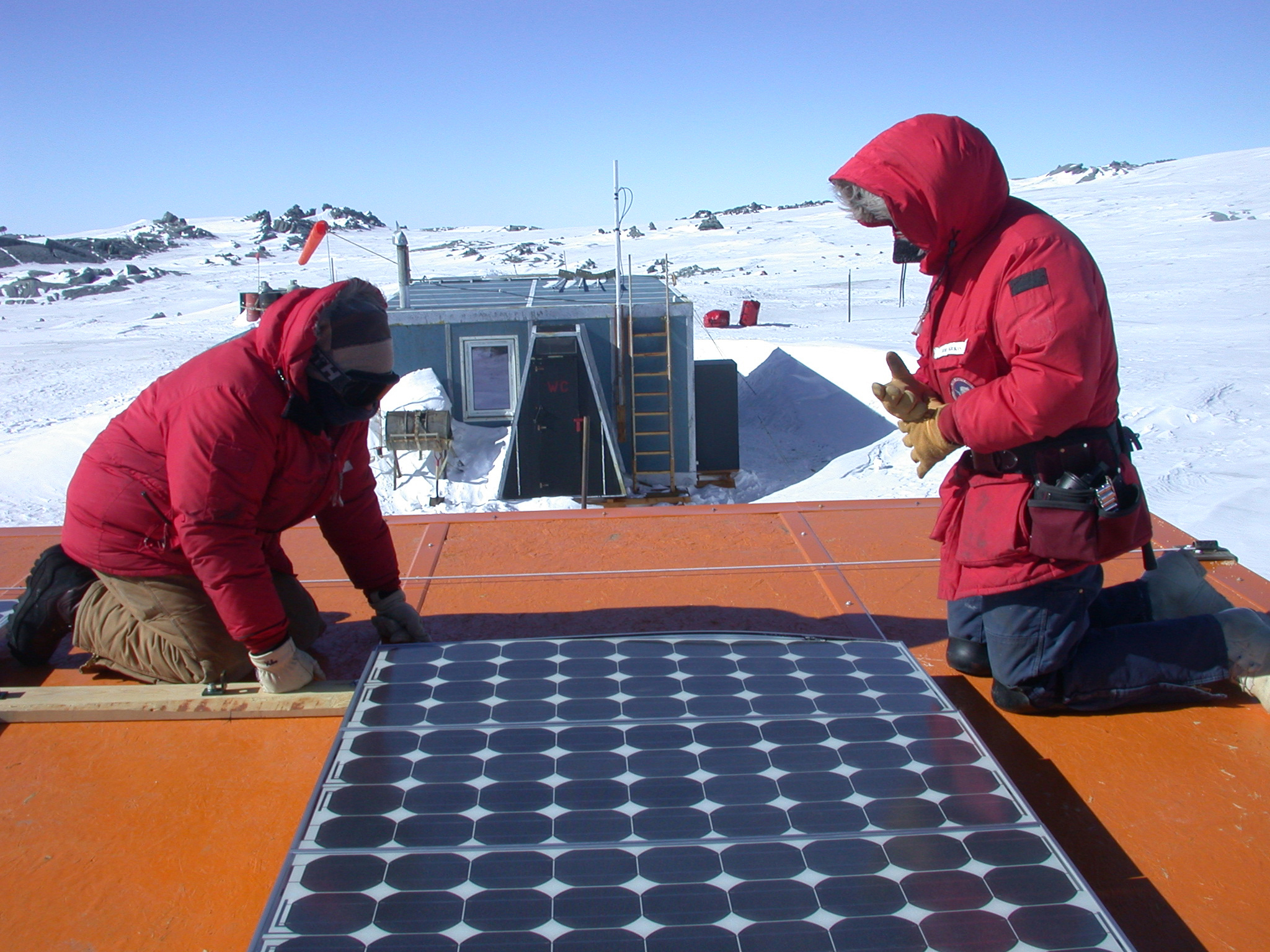 Two construction workers in red parkas kneel next to a solar panel while working outside.