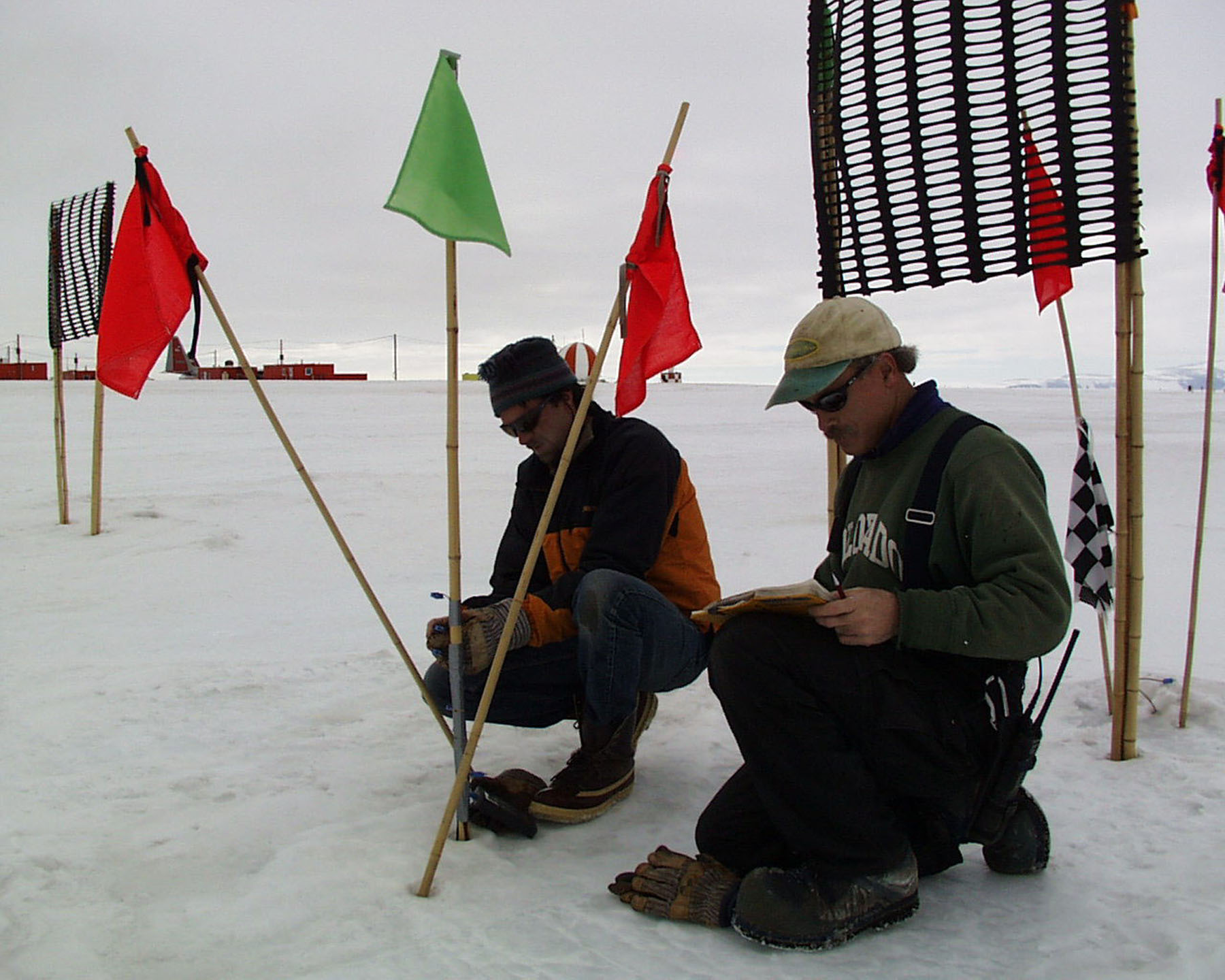 Two men kneel on the the ice near flagged bamboo poles.