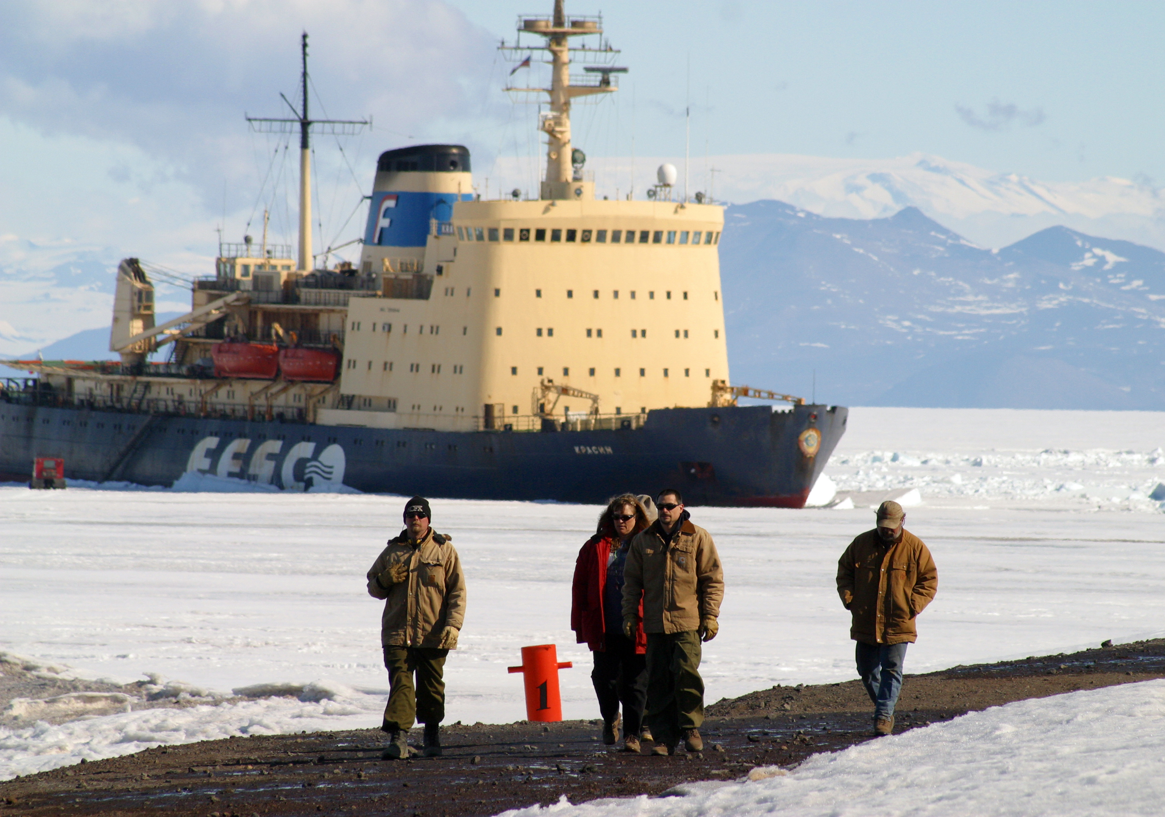 People walk on the shore in front of a boat in the ice.