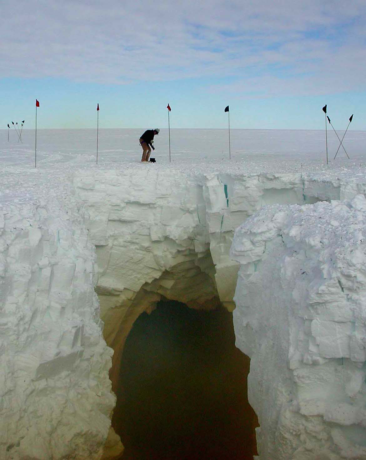A distant view of a person standing on a snow bridge over a large crevasse in the snow.