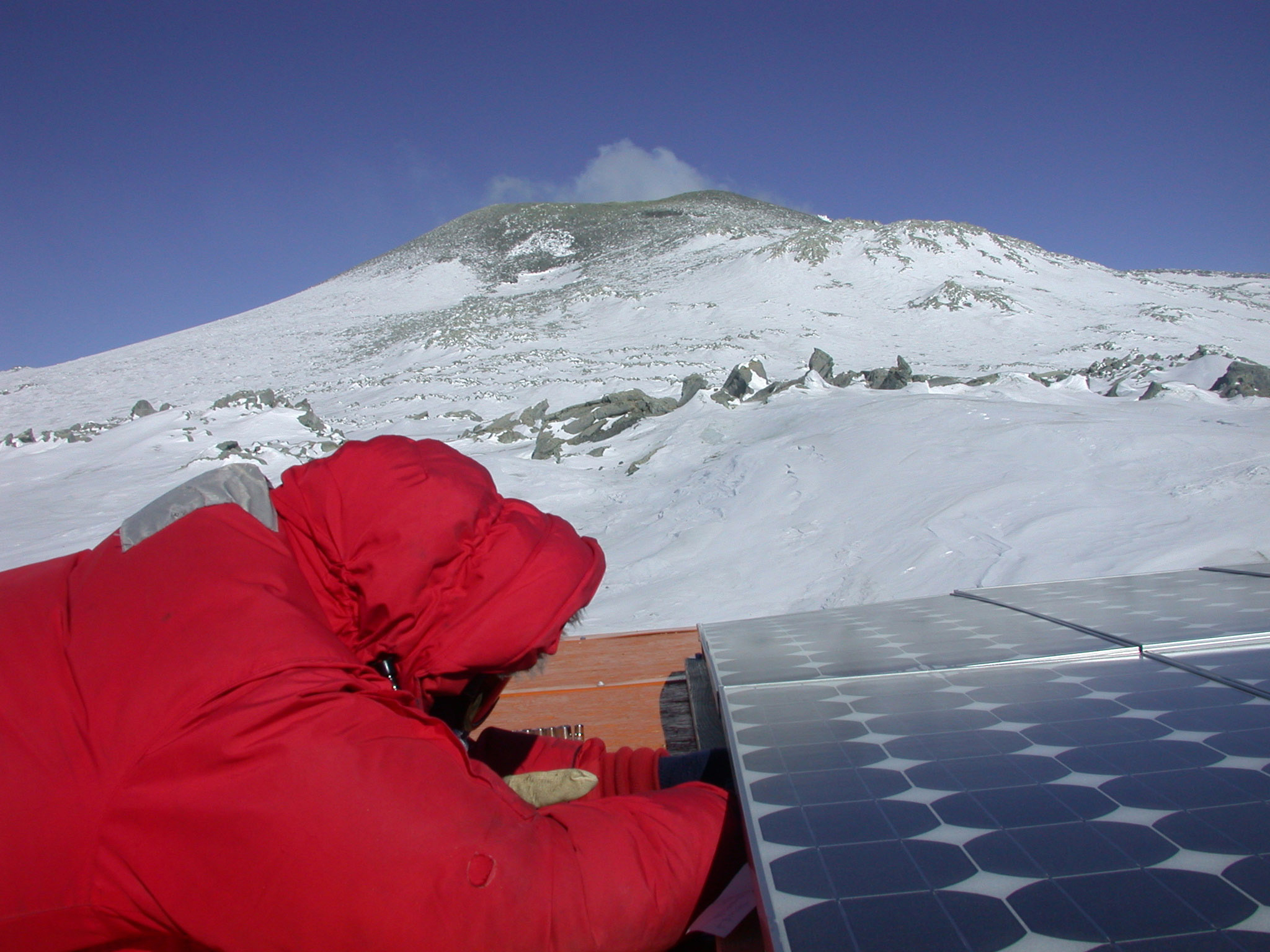 Man in a red parka works on a solar panel as a volcano belches smoke in the distance.