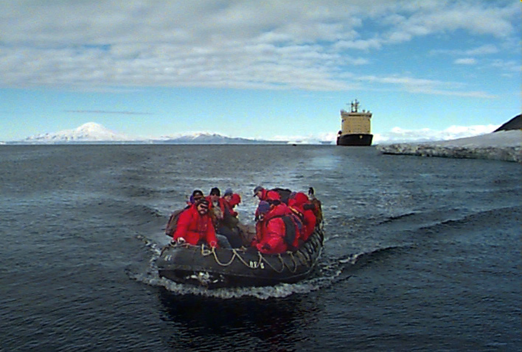 A group of people ride  in a rubber raft with a ship in the distance.
