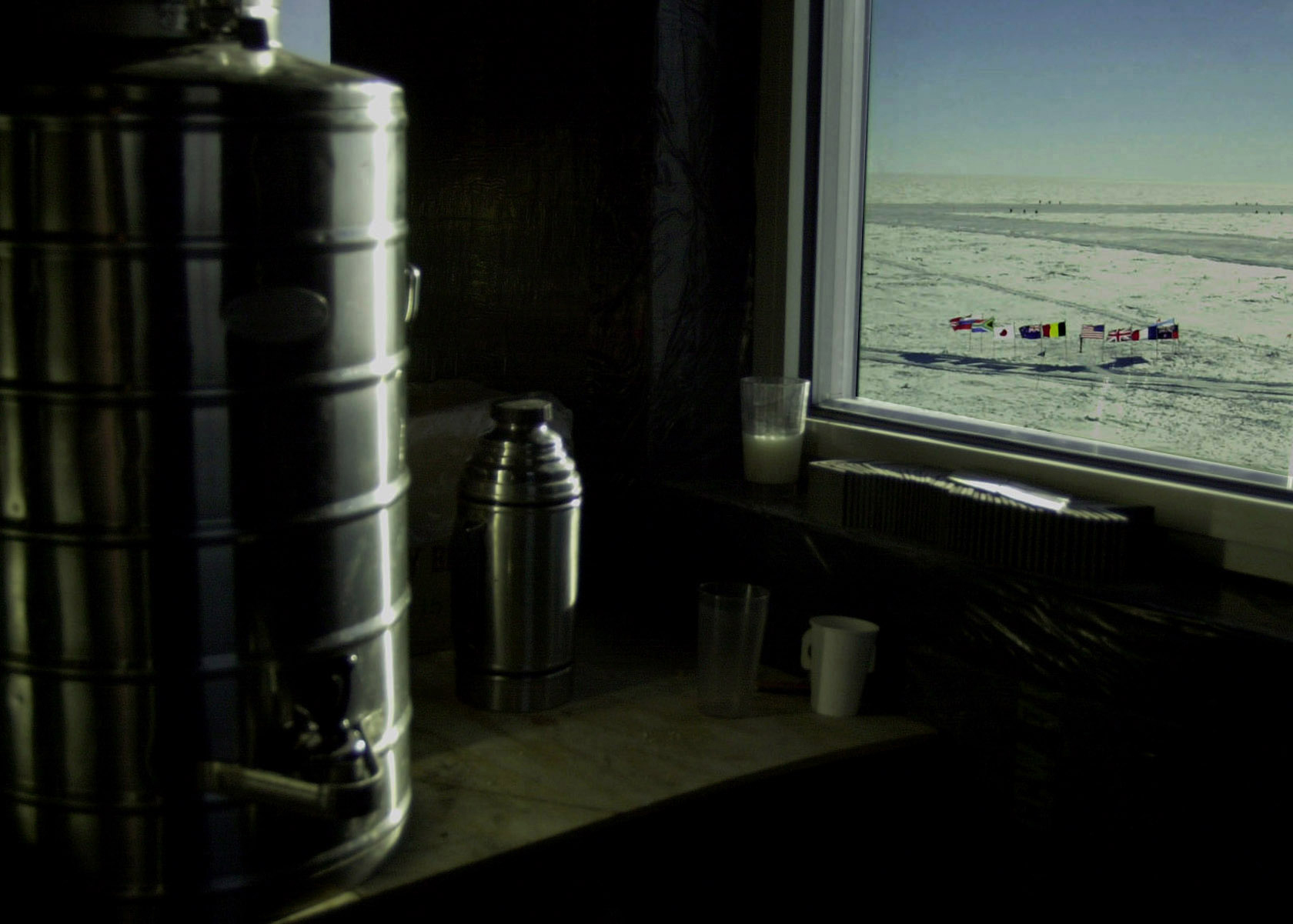 Interior shot of coffee thermos near window with view out the window of many national flags fluttering in the wind.  The distant landscape is white snow.