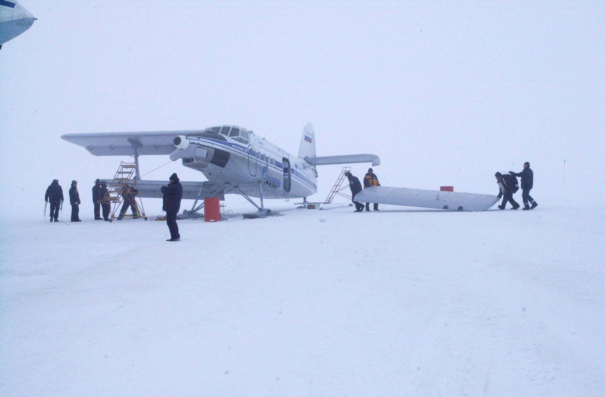 People working near a biplane on snow, with one set of wings removed.