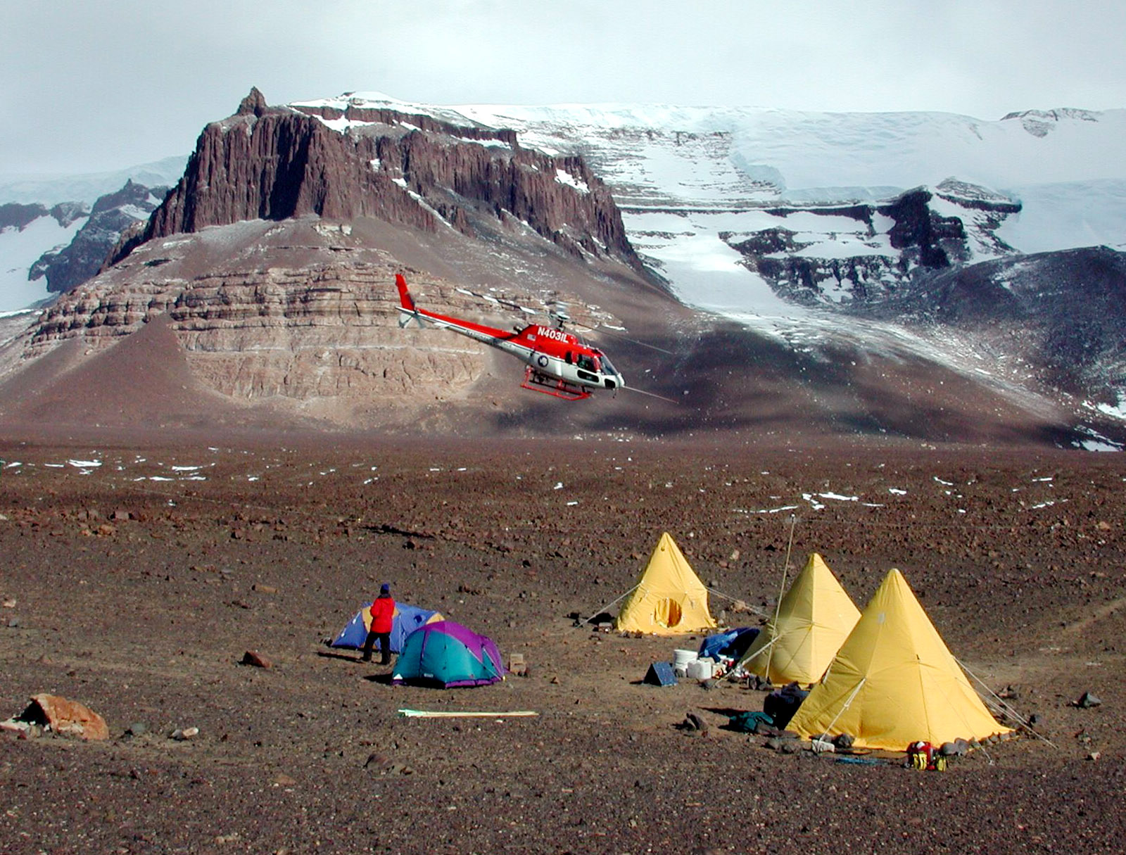Helicopter approaches several tents on a rocky landscape.