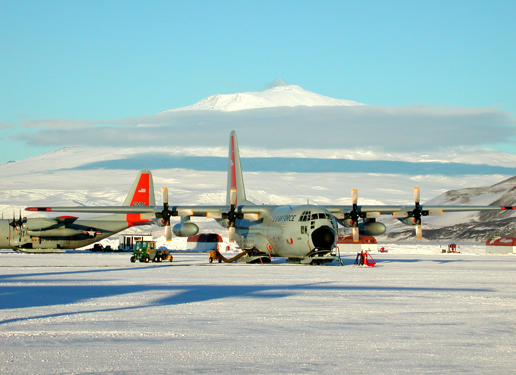 Airplanes with four propellers and skis, sit on ice, with a snow-covered mountain in the distance.