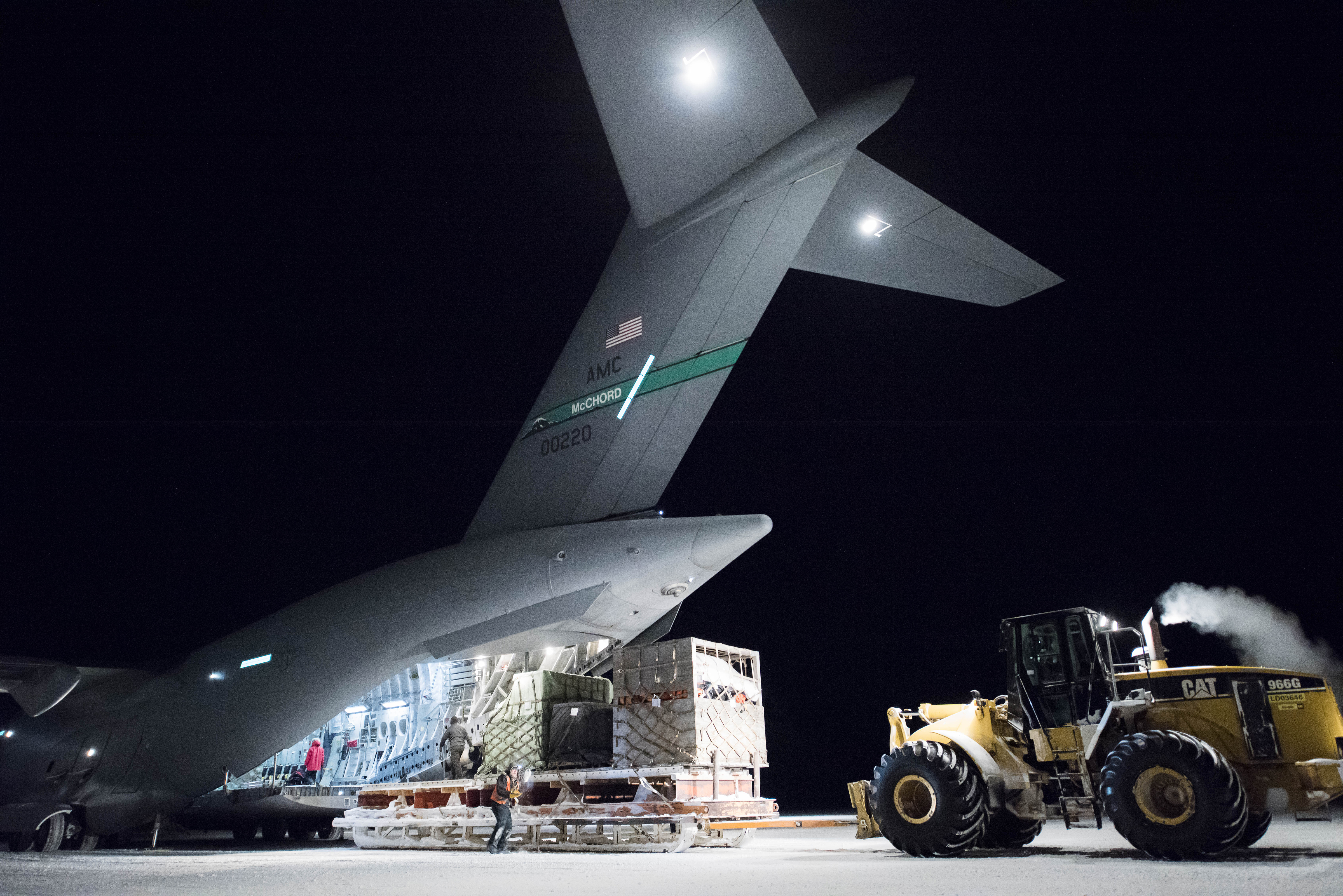 A forklift unloads a cargo pallet from the rear of a large Air Force cargo jet during the night.