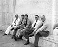 Five men in uniforms and neckties sit informally against a marble building.