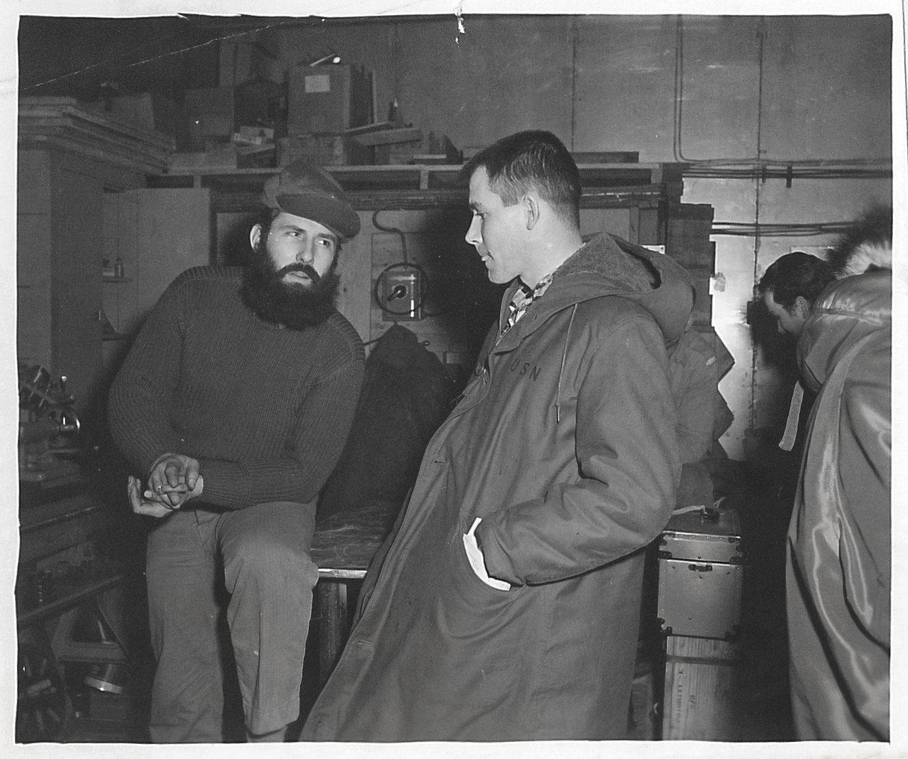 Two men talking in a room. Others in the background.