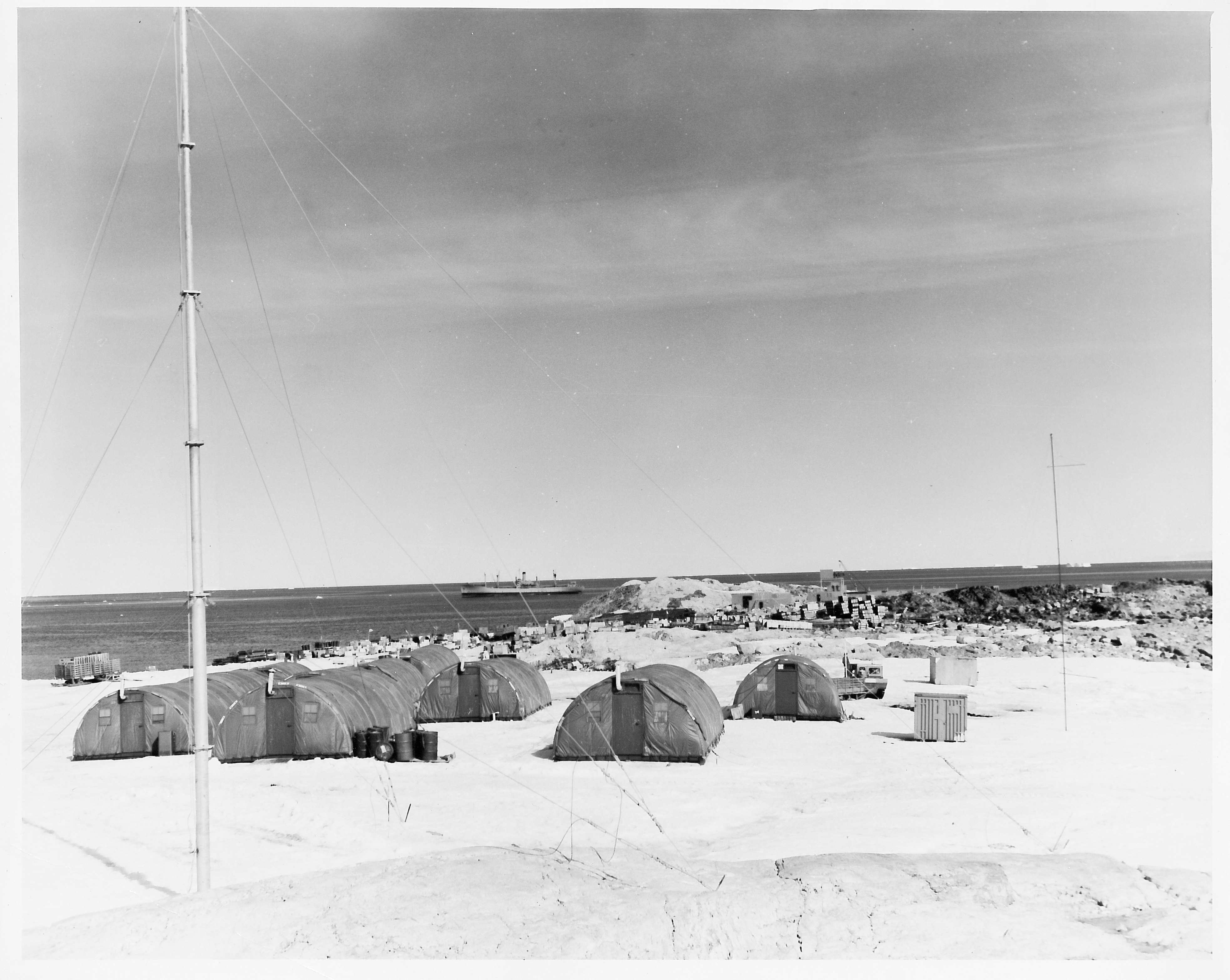 A camp consisting of large tents, water and a ship far in the distance.