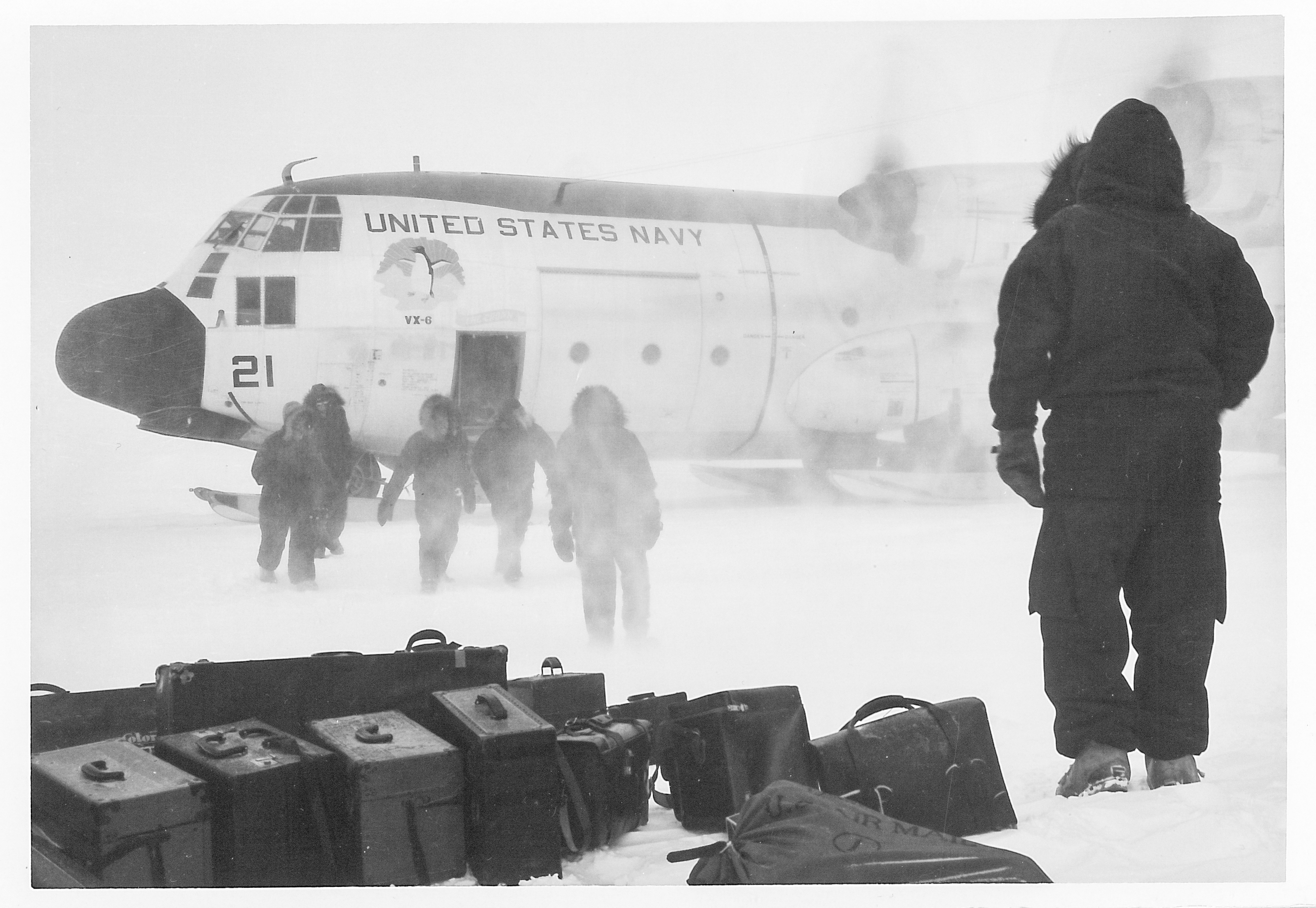 A man standing next to cargo as other men exit an airplane.