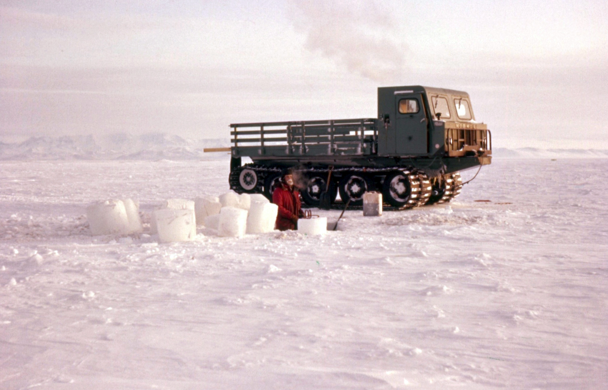 A man digging a hole in ice, with a tracked vehicle parked nearby.