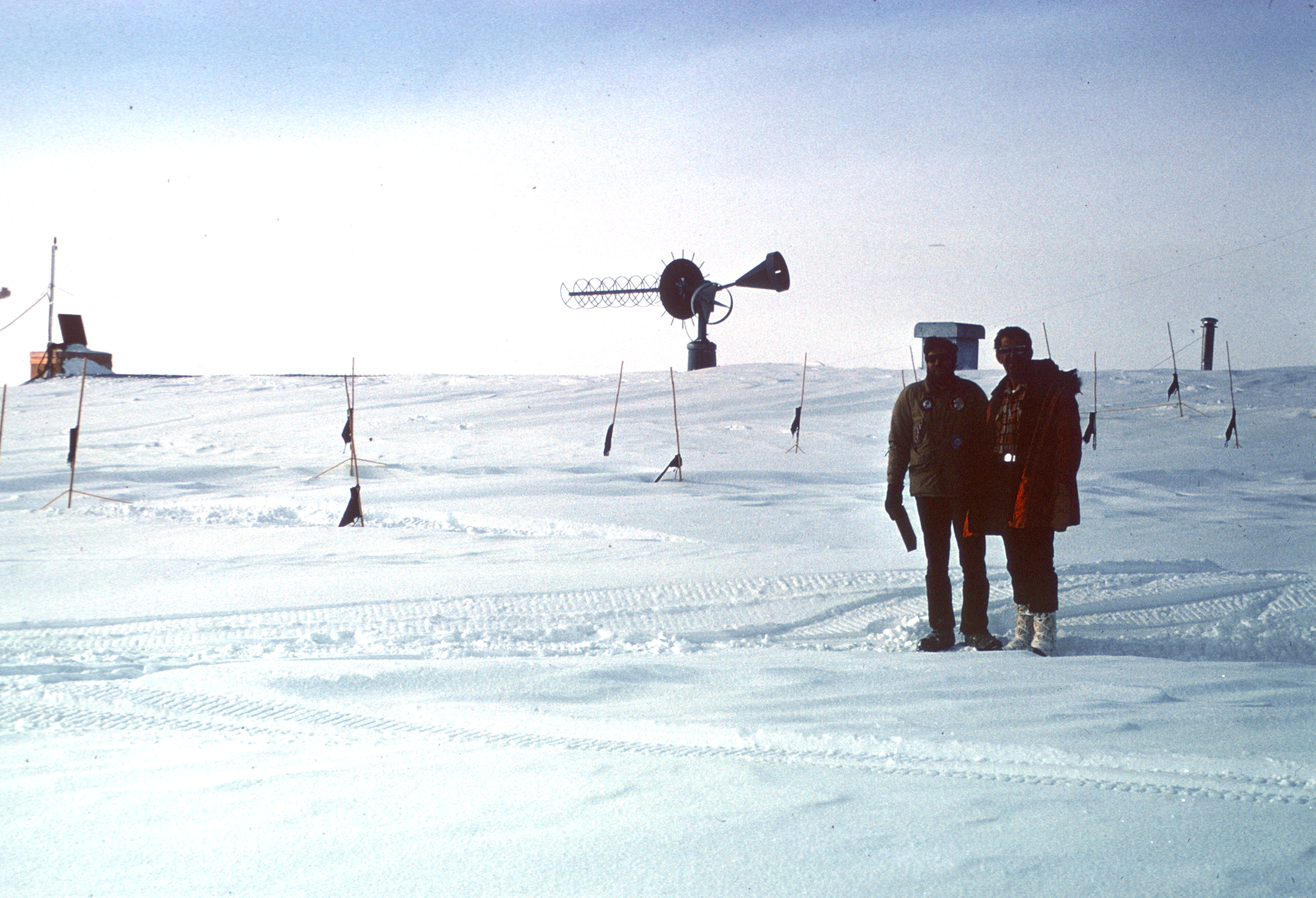 Two men stand on a snowy field with a large scientific instrument behind them.