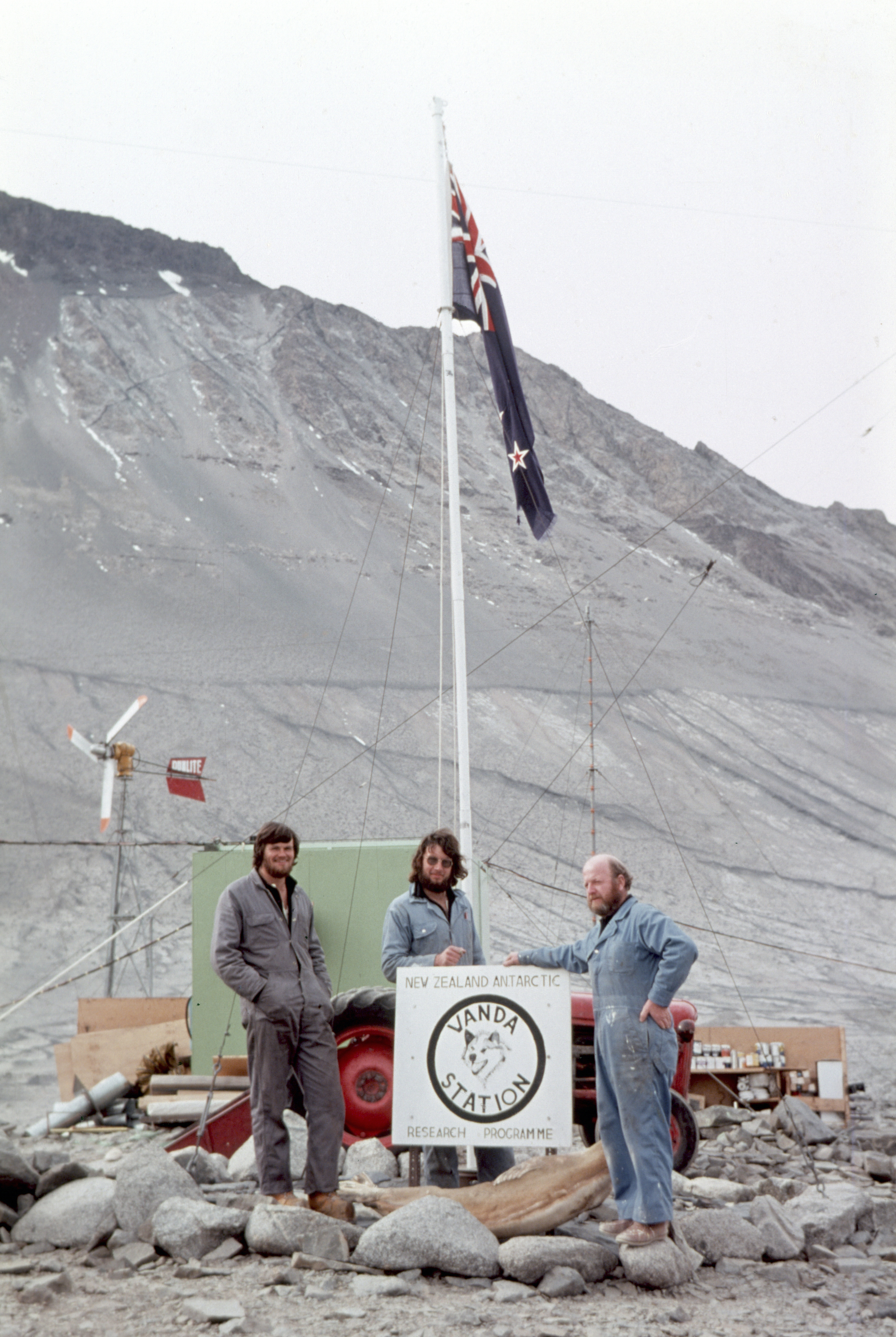 Three men stand next to a sign and flag pole in a rocky valley.