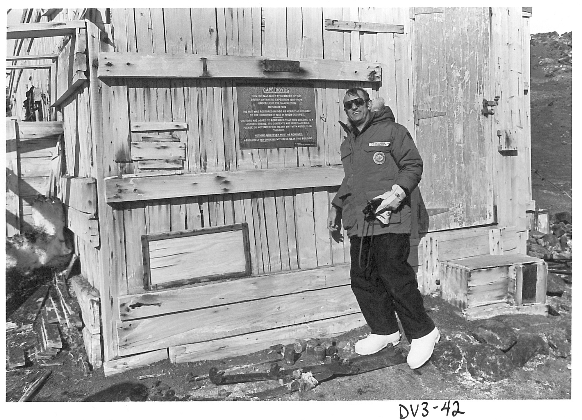 Wearing cold-weather gear a man stands outside a wooden building.