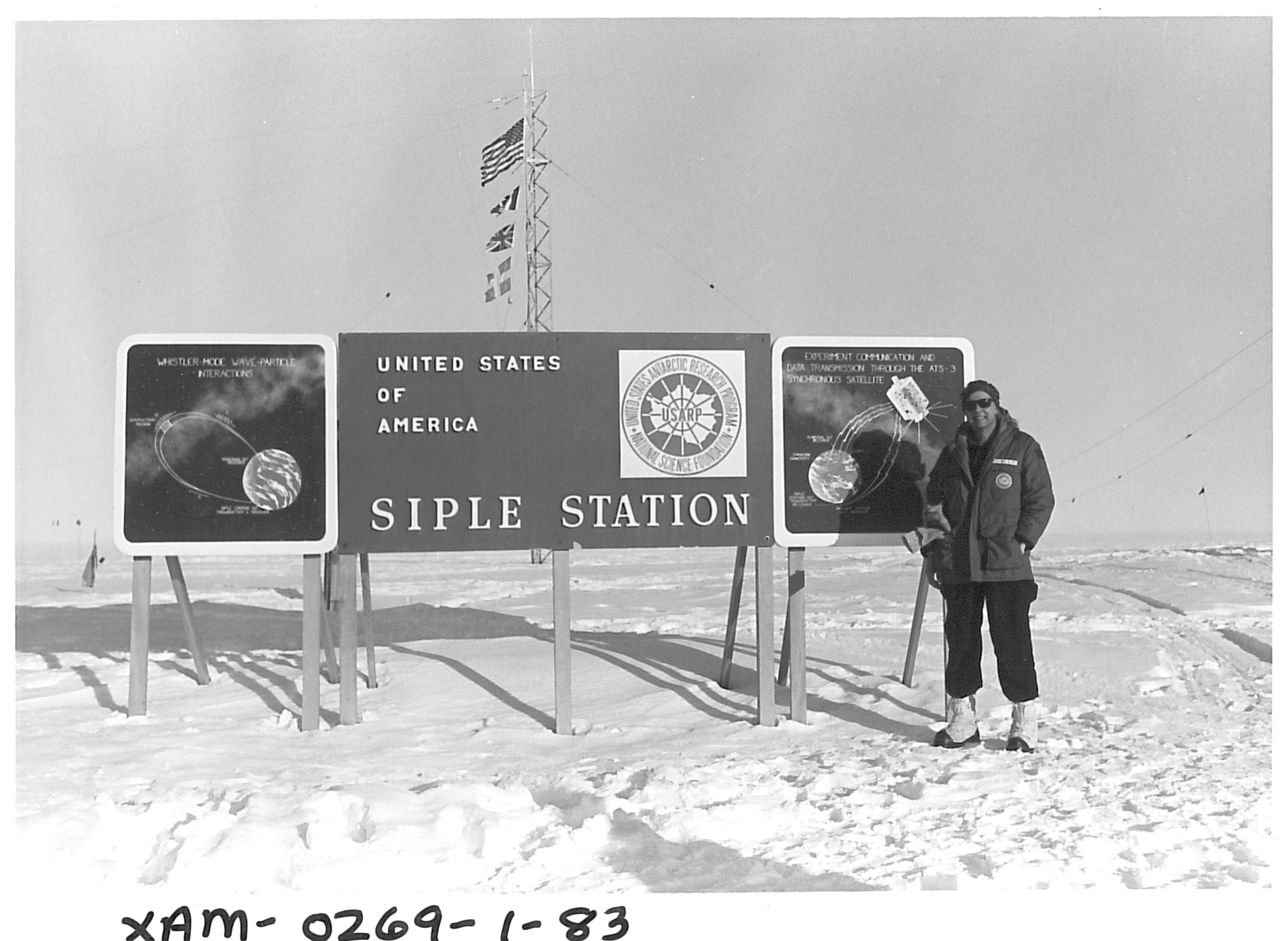 A man standing on snow-covered ground next to a sign and a flagpole. 