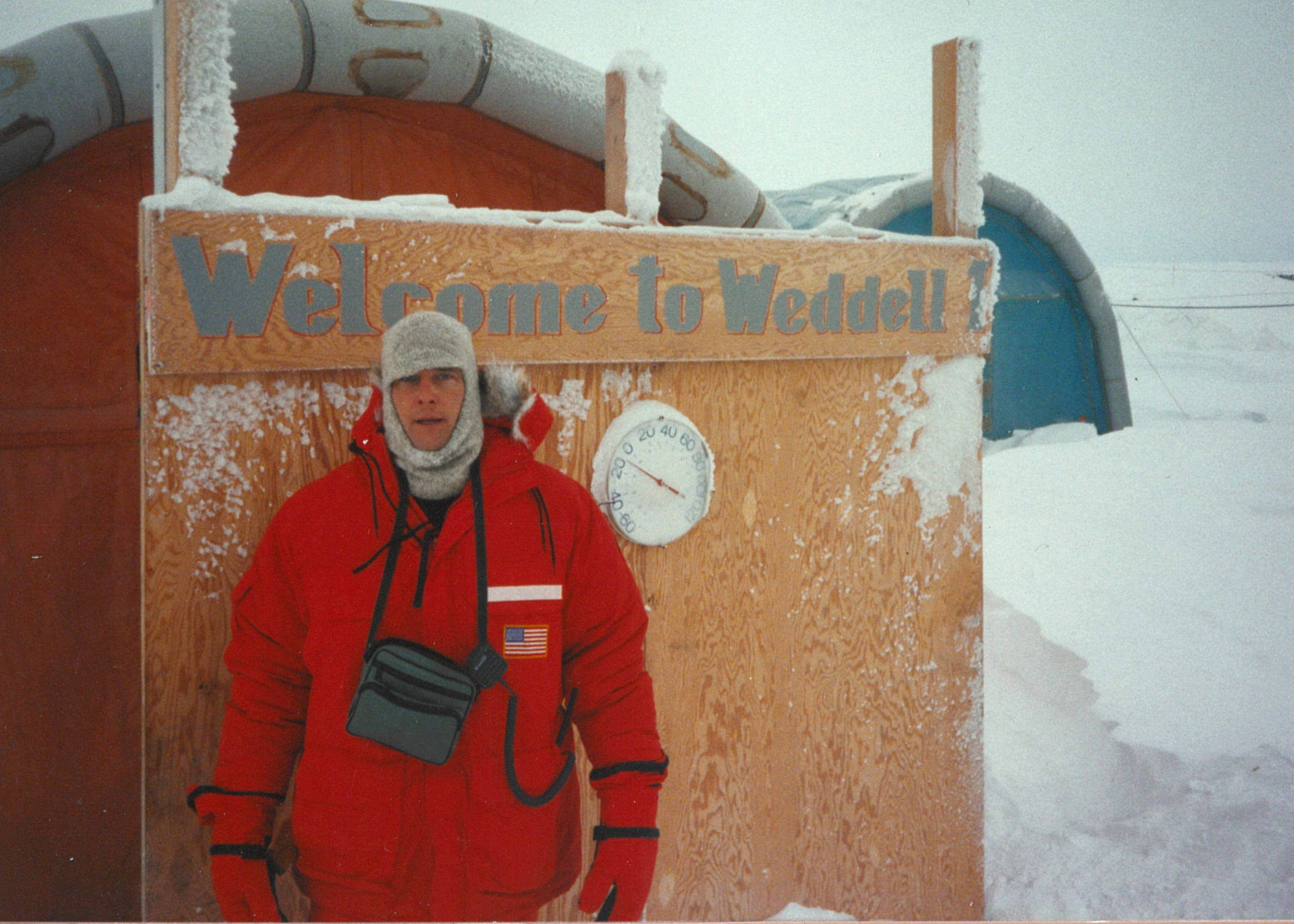 A man in a red parka in front of a sign that says 'Welcome to Weddell.' A thermometer indicates a temperature of about -10°F.