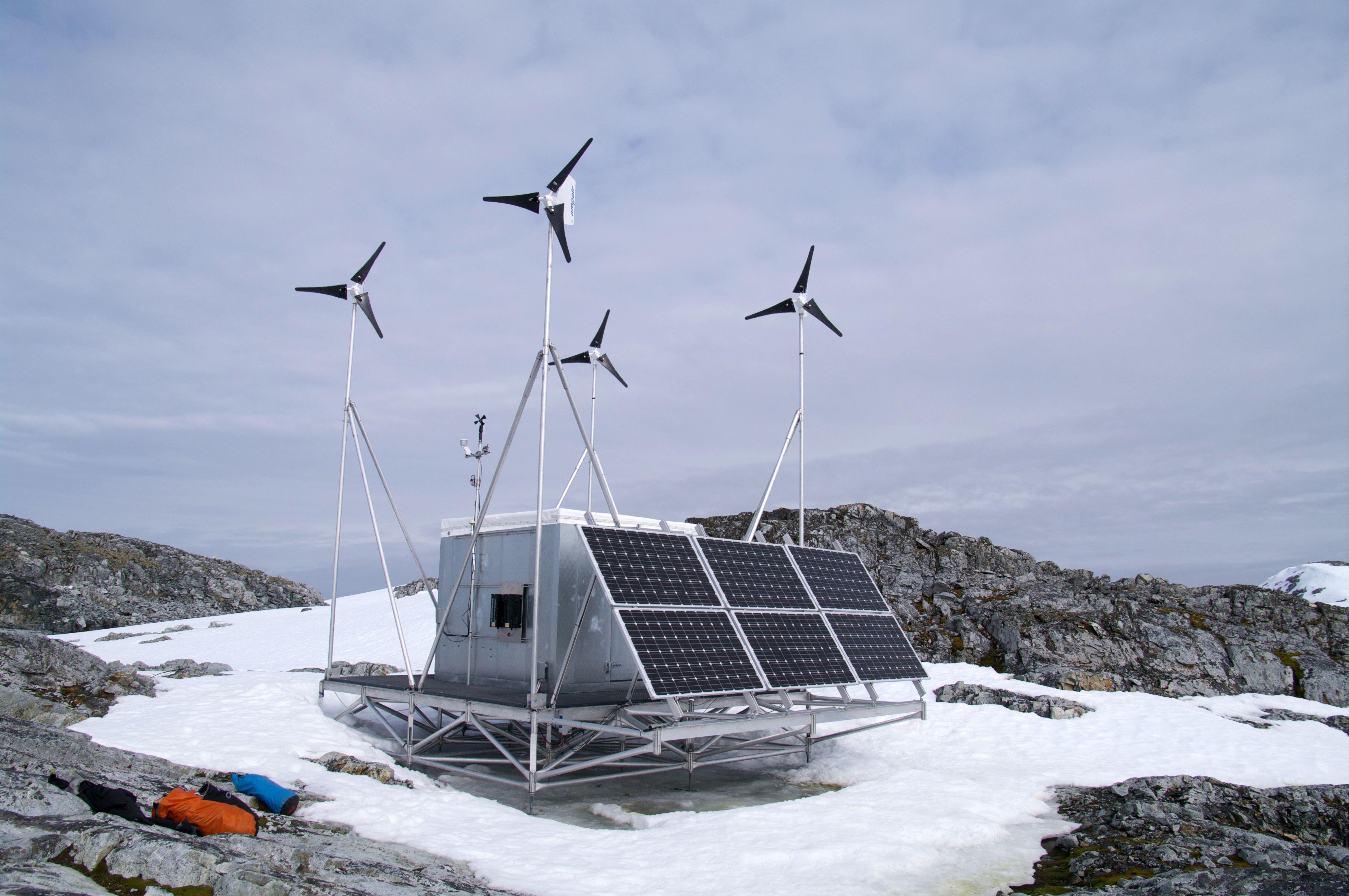 A hut with solar panels and wind turbines sits in a snow patch on rocky ground.