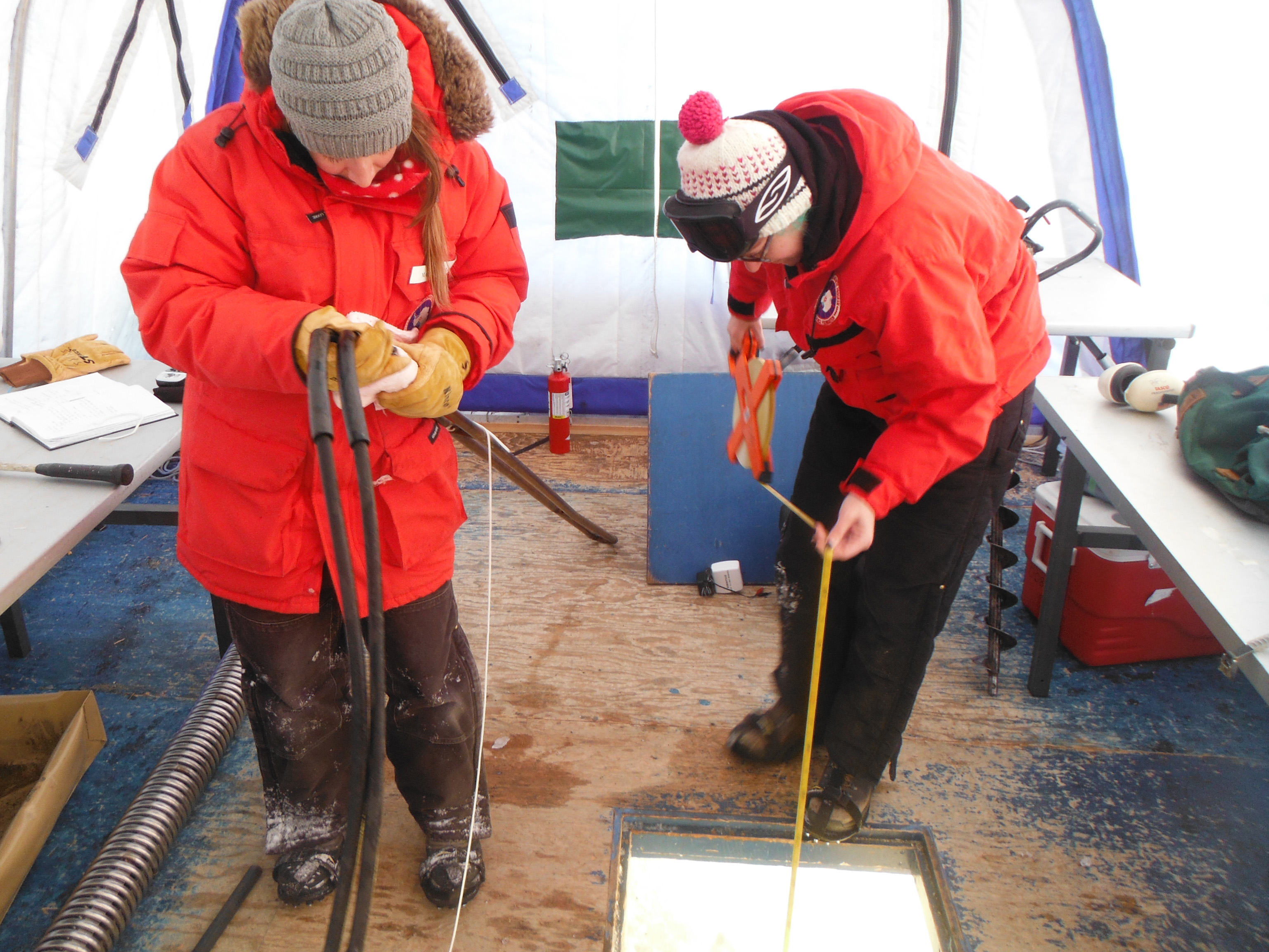 Two people in red coats working over a hole in the floor of a tent shelter.