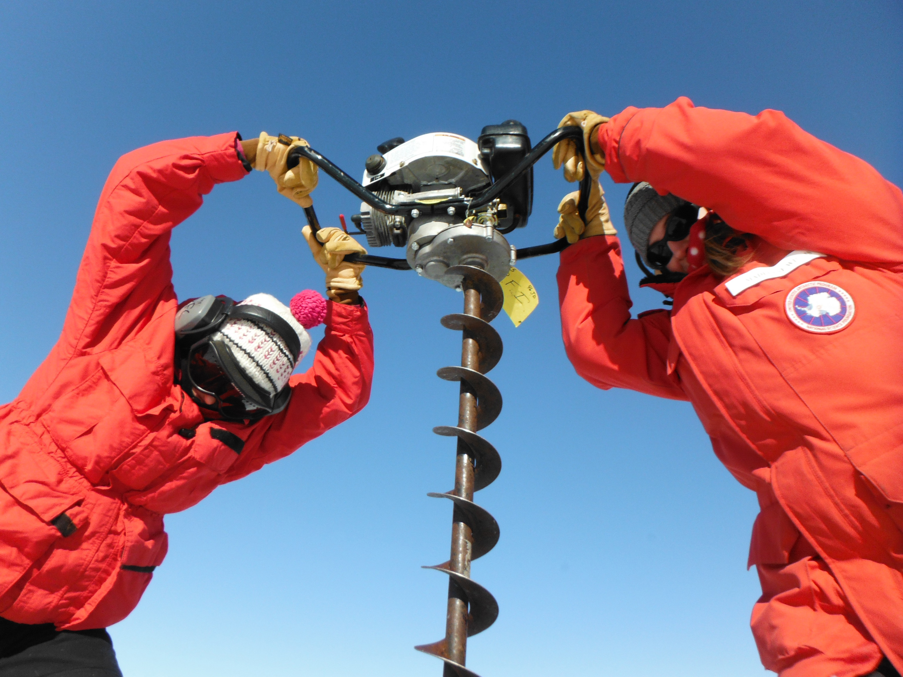 Two people in red coats use a motorized auger.