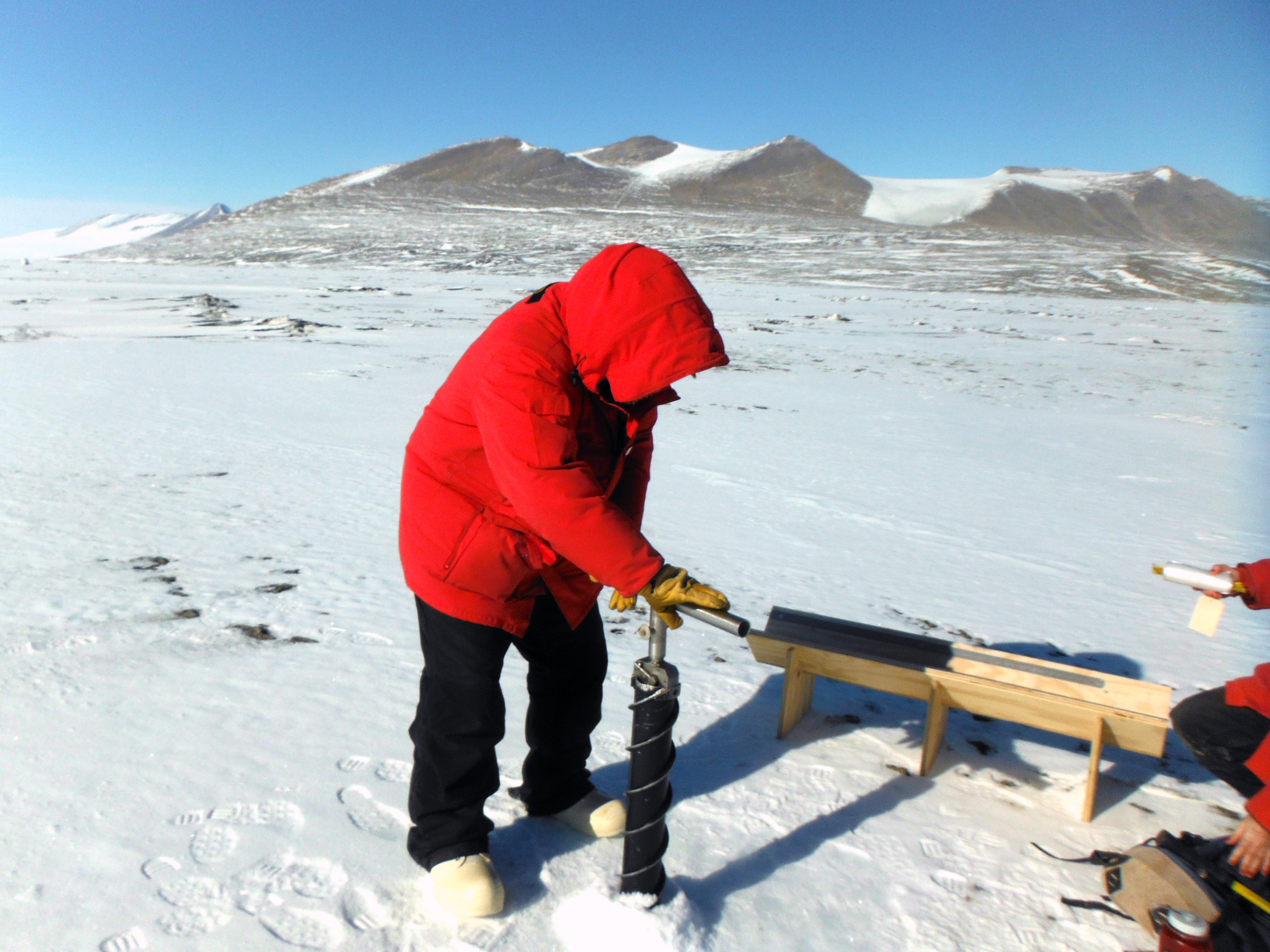 A person in a red parka uses a hand auger to drill down into surface ice.