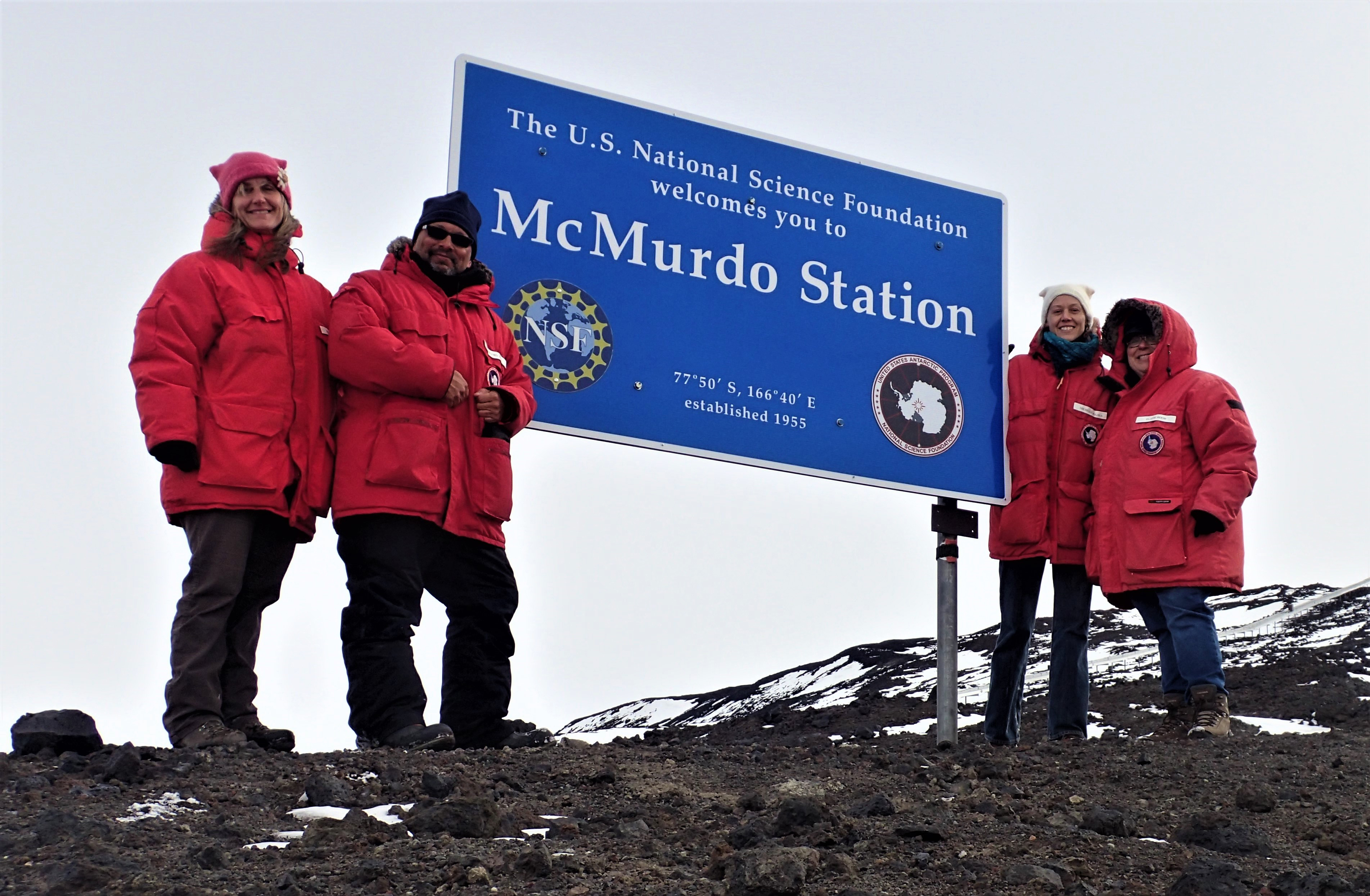 Four people in red winter parkas stand next to a large blue sign.
