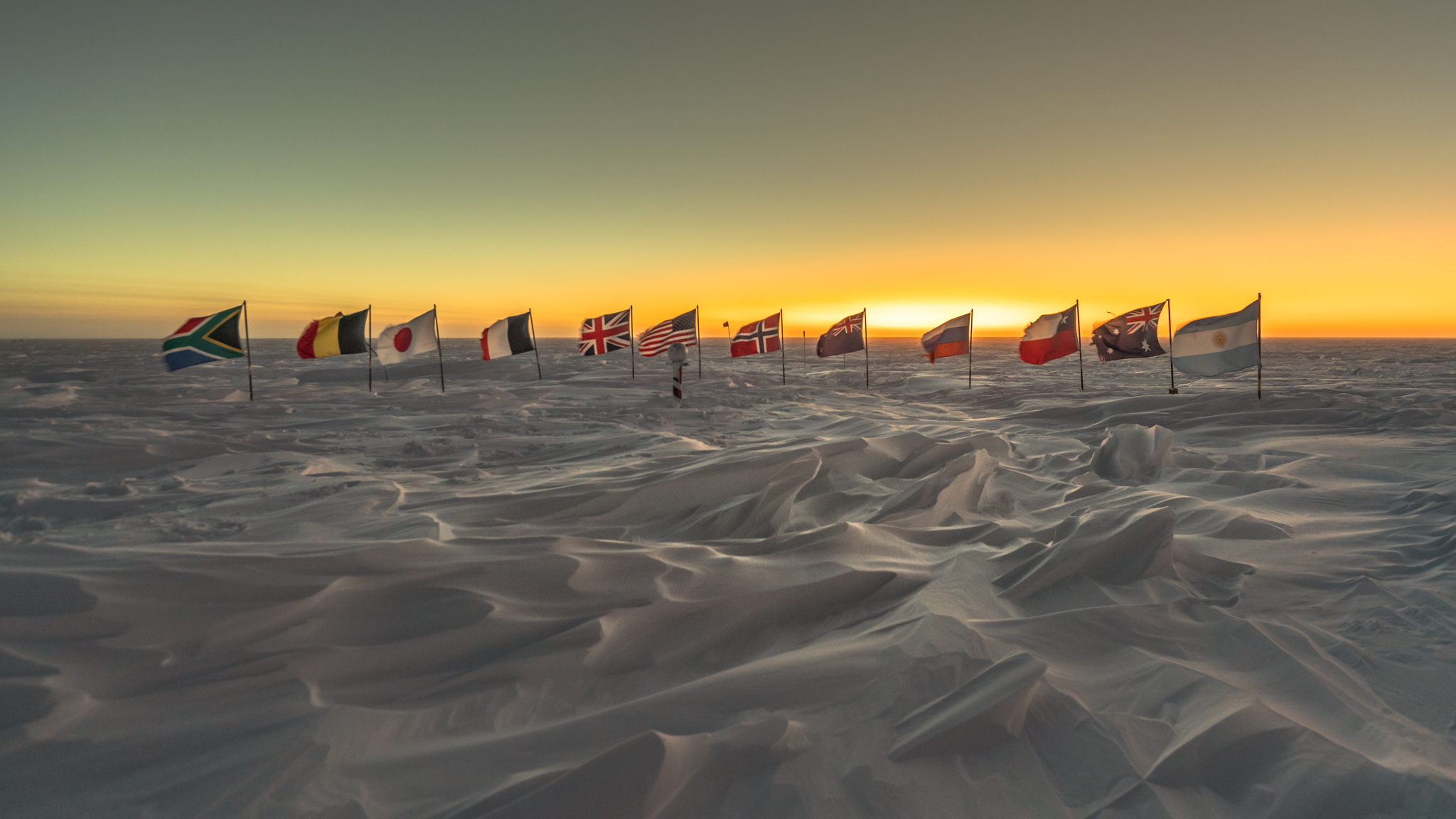 Twelve countries' flags in the snow with the sunrise behind them.