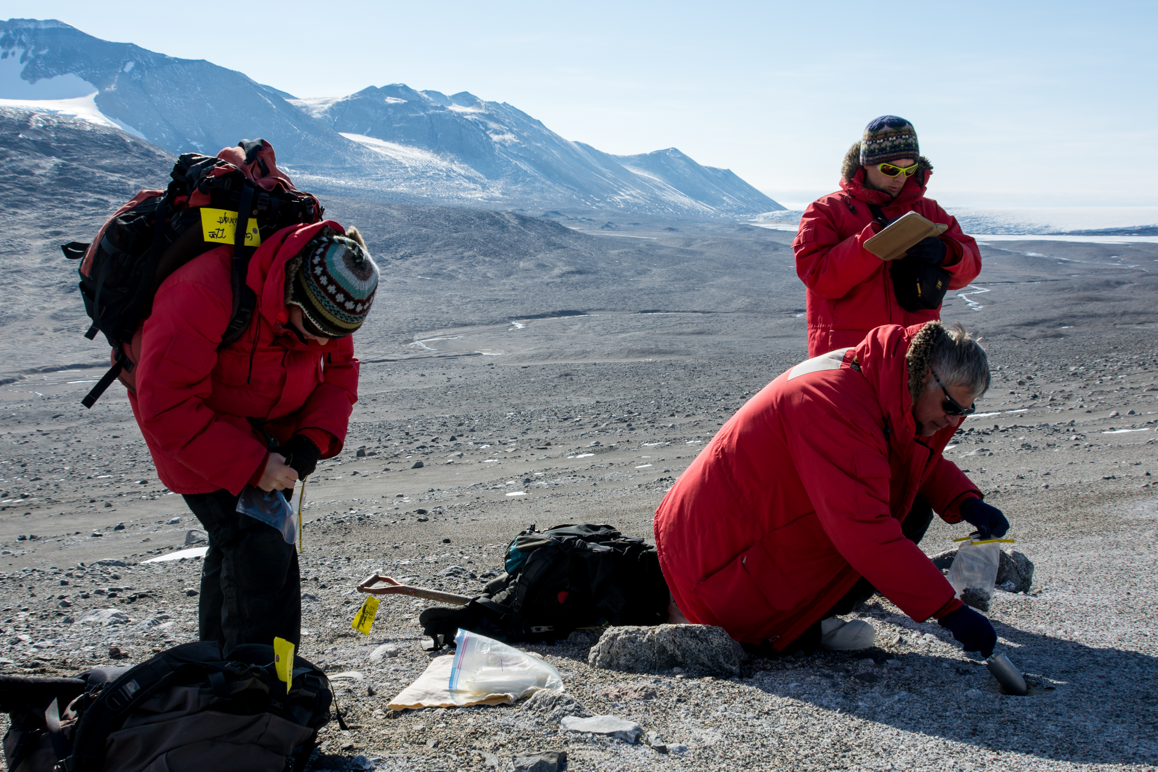 Three people in red parkas gathering soil samples.