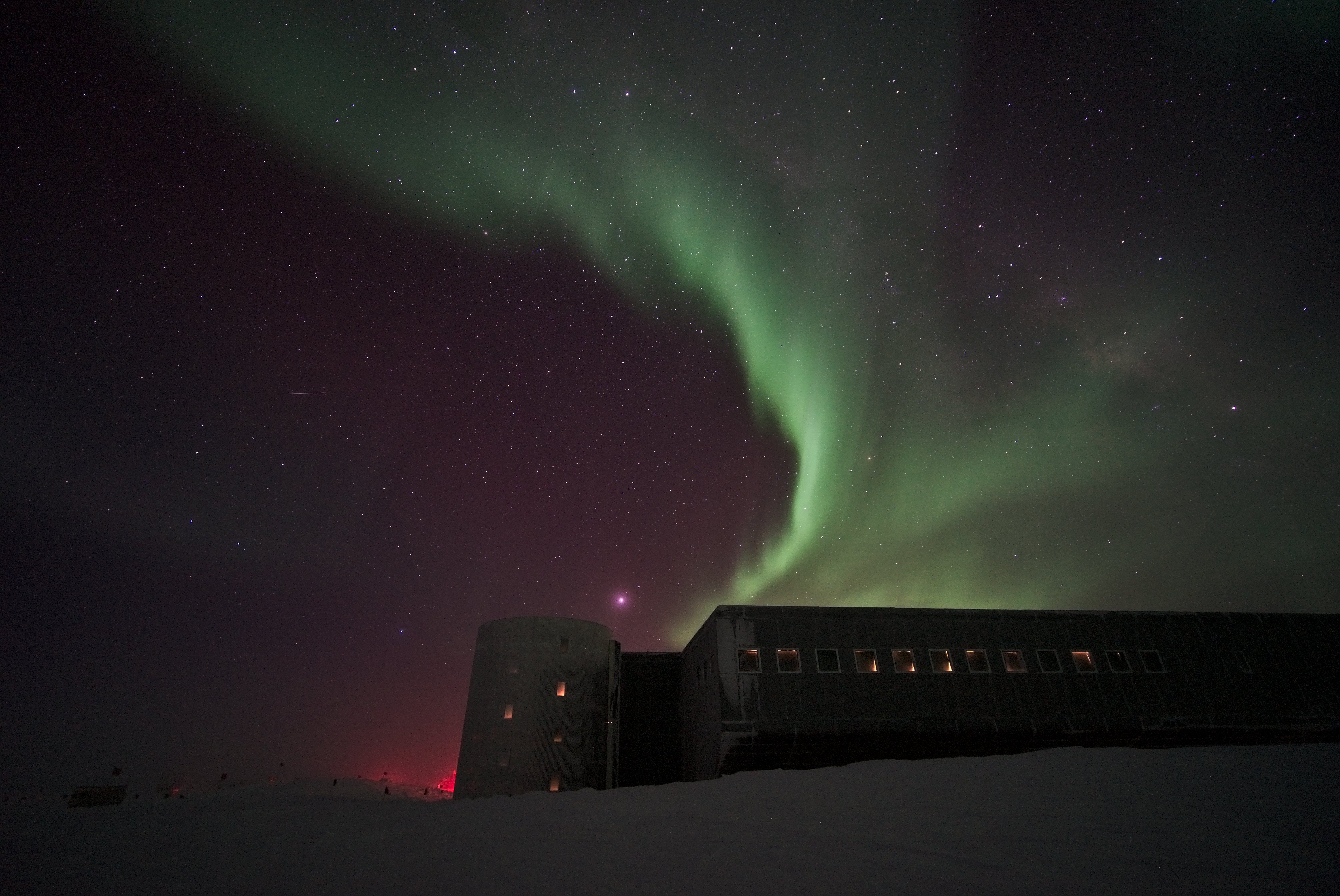 Green auroras over a building and snow, with the night sky beyond. 