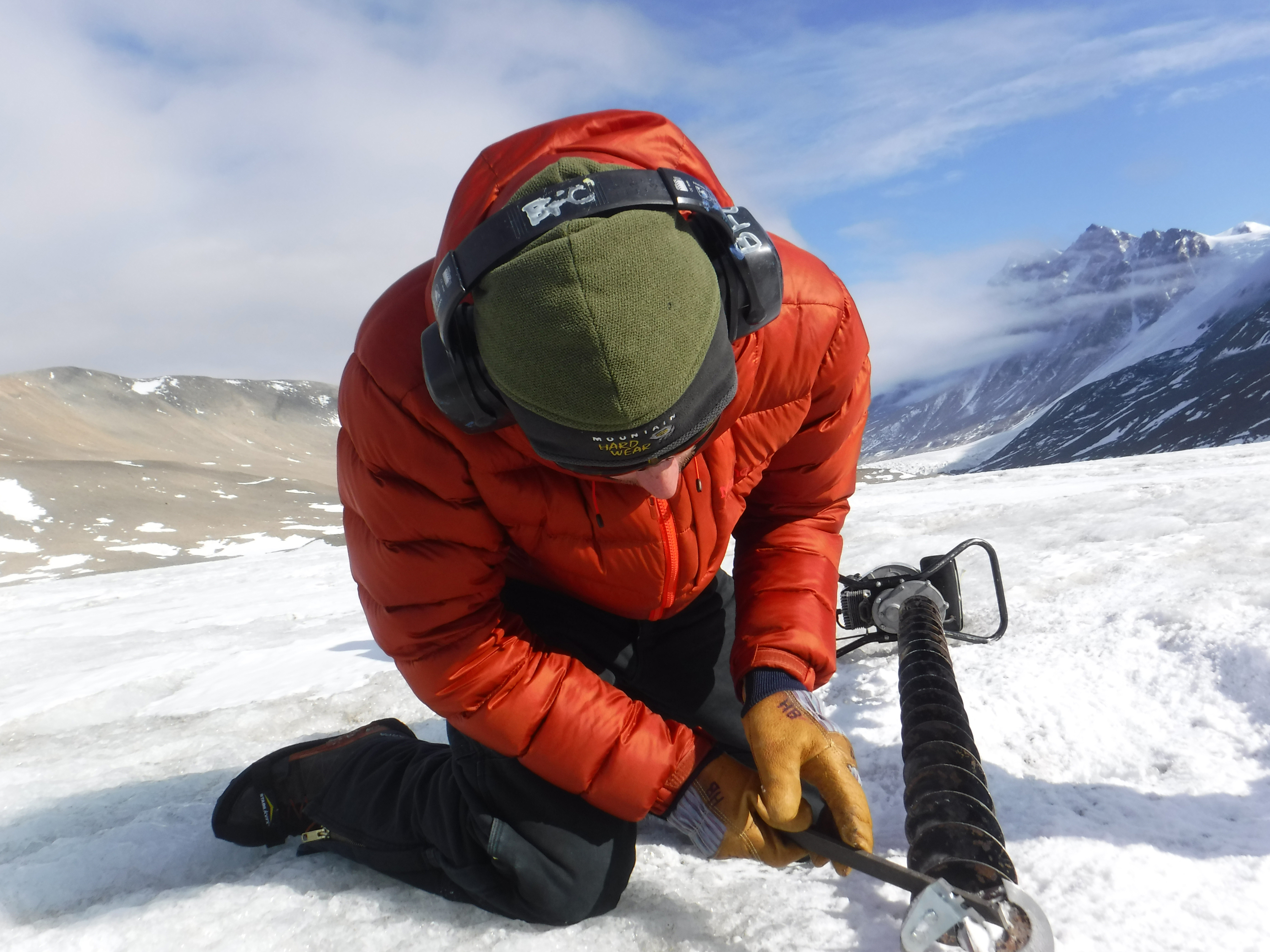 A person kneels on snow as he works with a motorized auger.