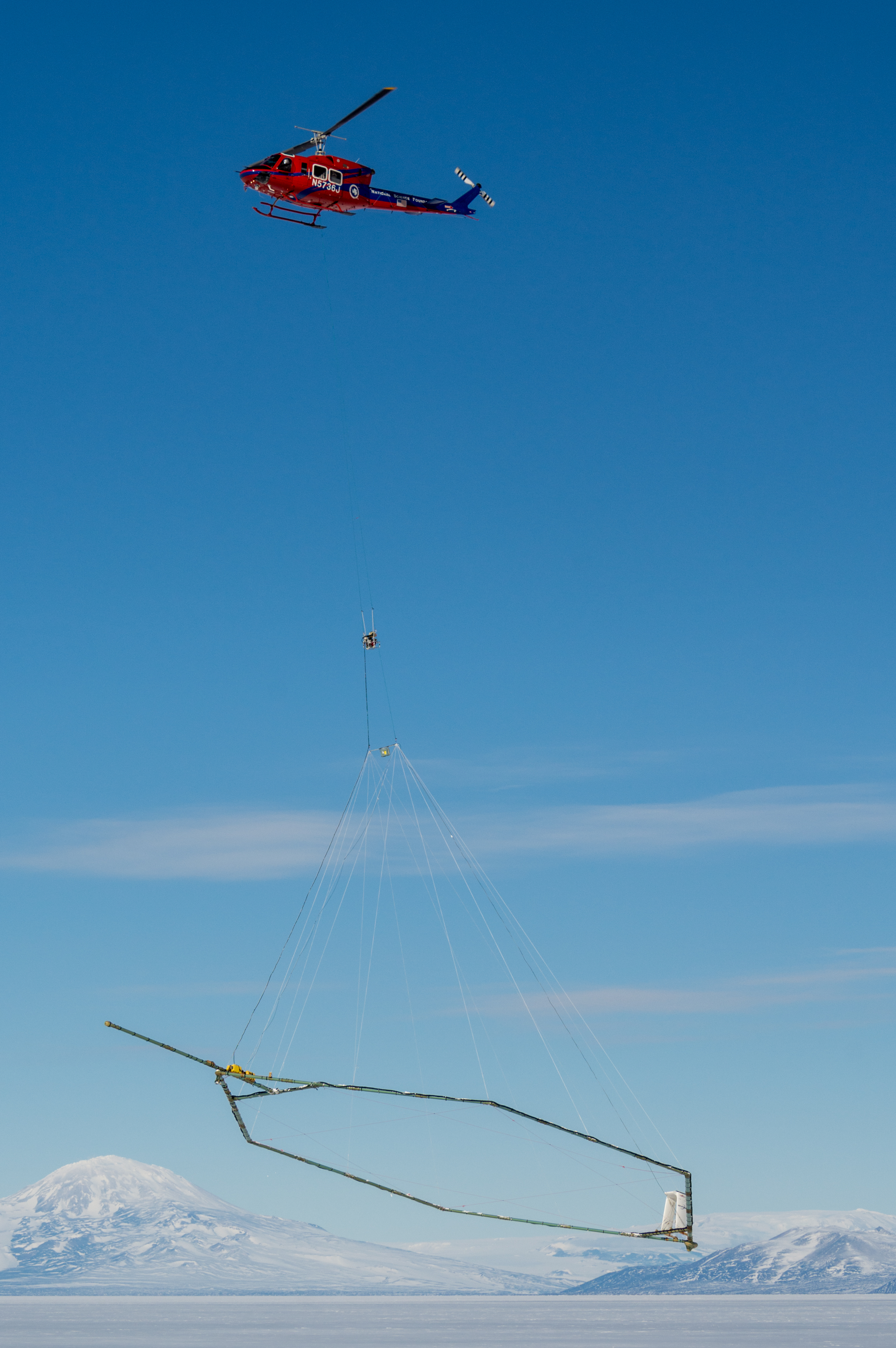 A helicopter with a scientific instrument under it takes off from a sheet of ice with mountains in the background. 