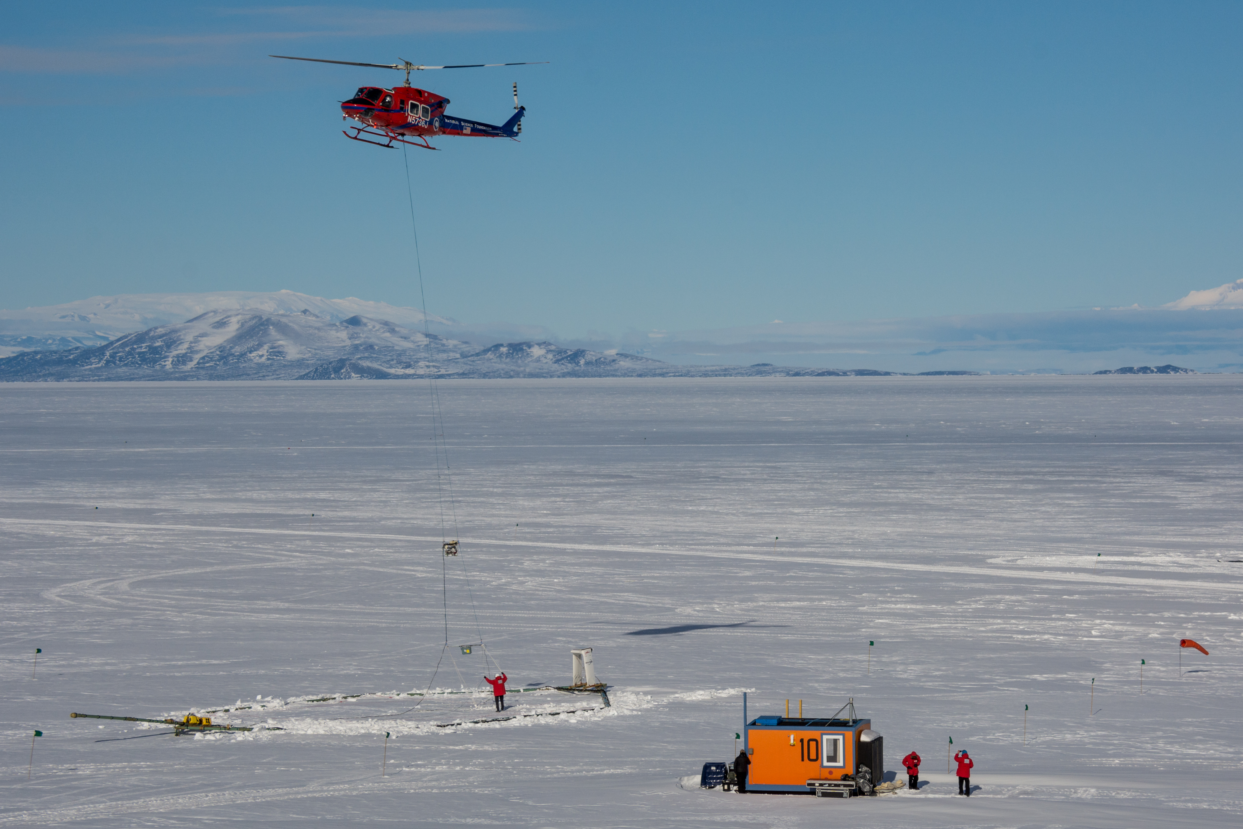 A helicopter takes off from a sheet of ice with a large instrument slung below it.