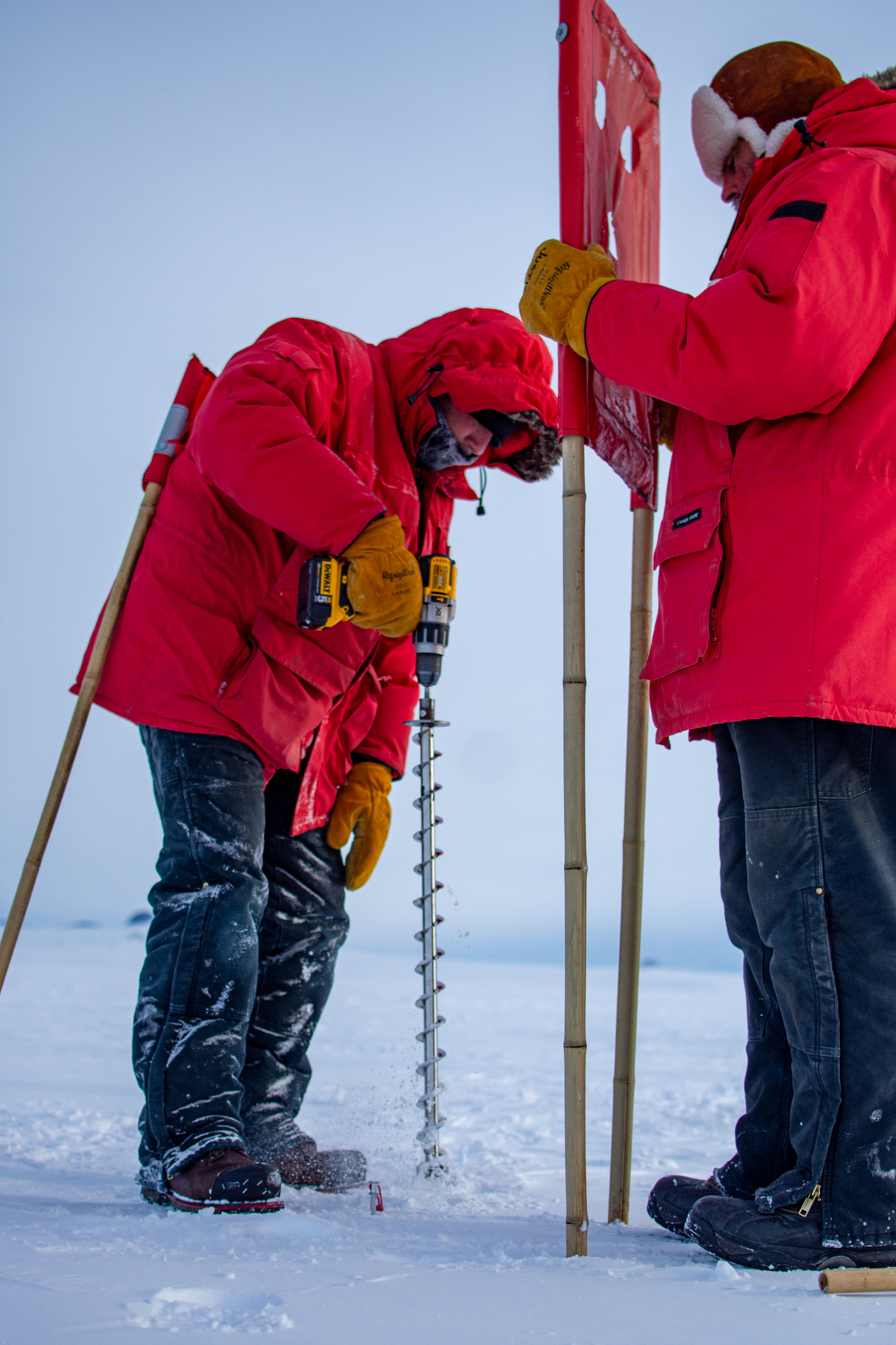 One person drills a hole in the ice while a second person holds an airfield marker.