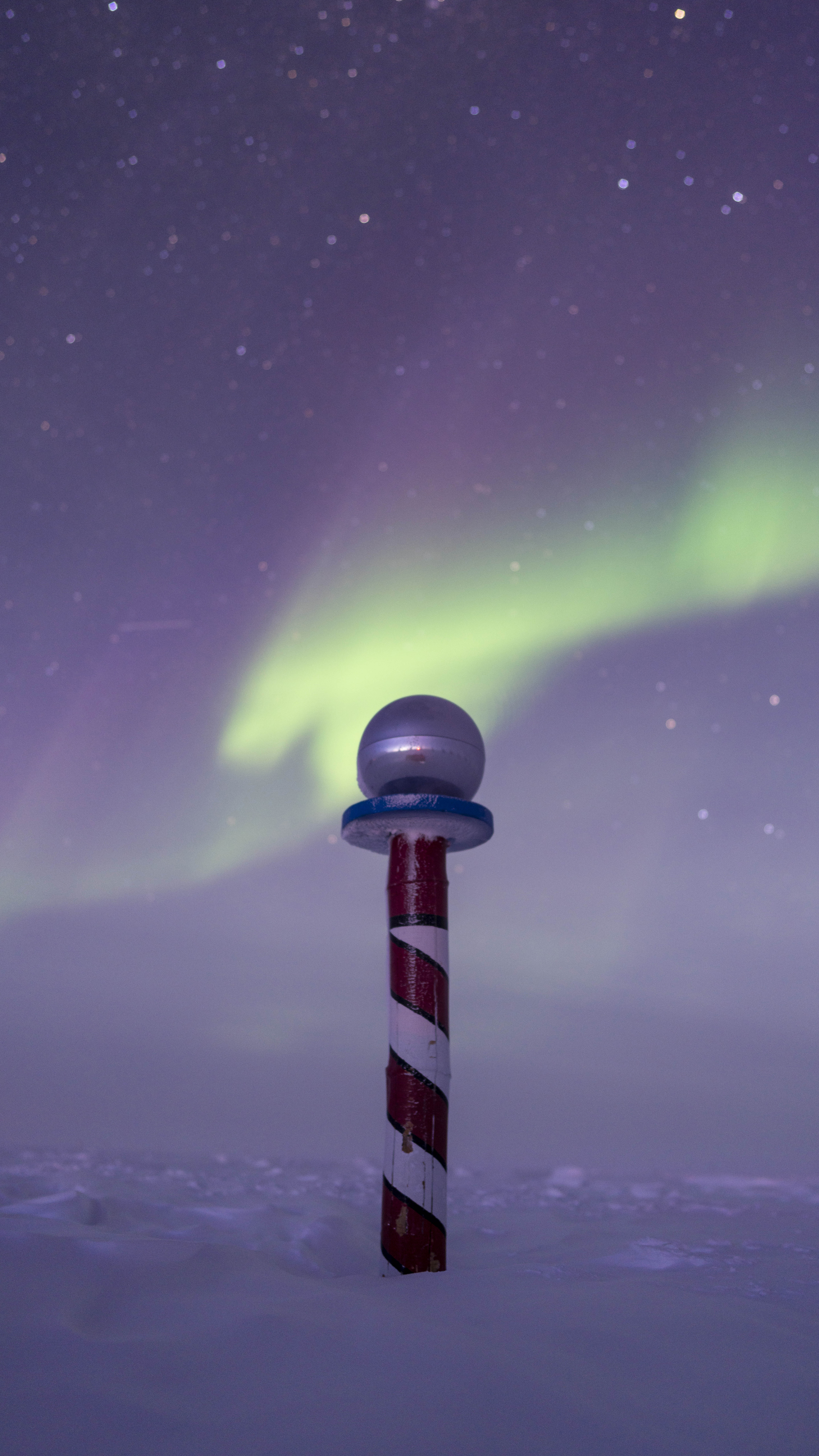 Auroras fill the sky behind a red and white striped pole.