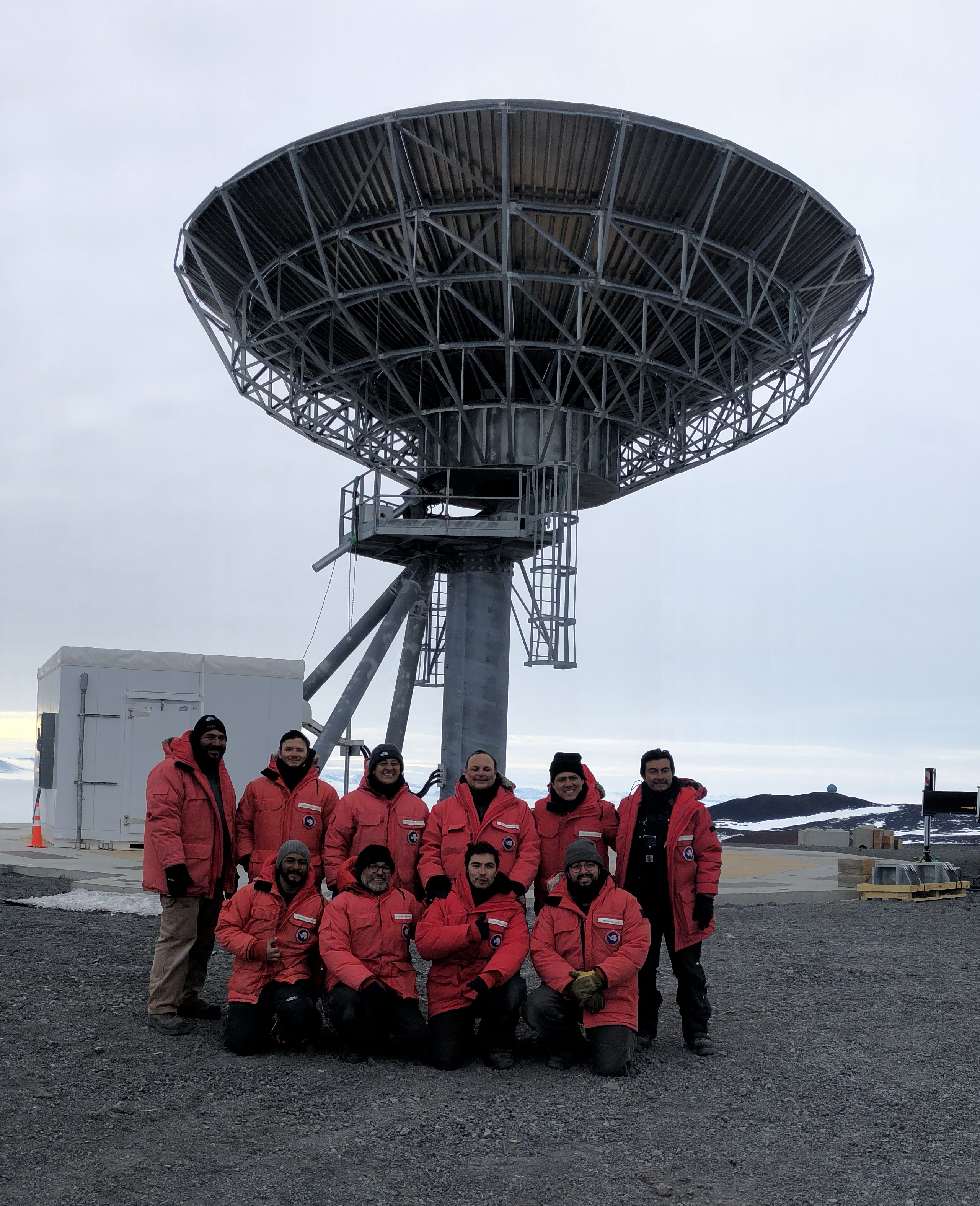 Ten men in red parkas pose for a photo in front of a satellite communications dish.