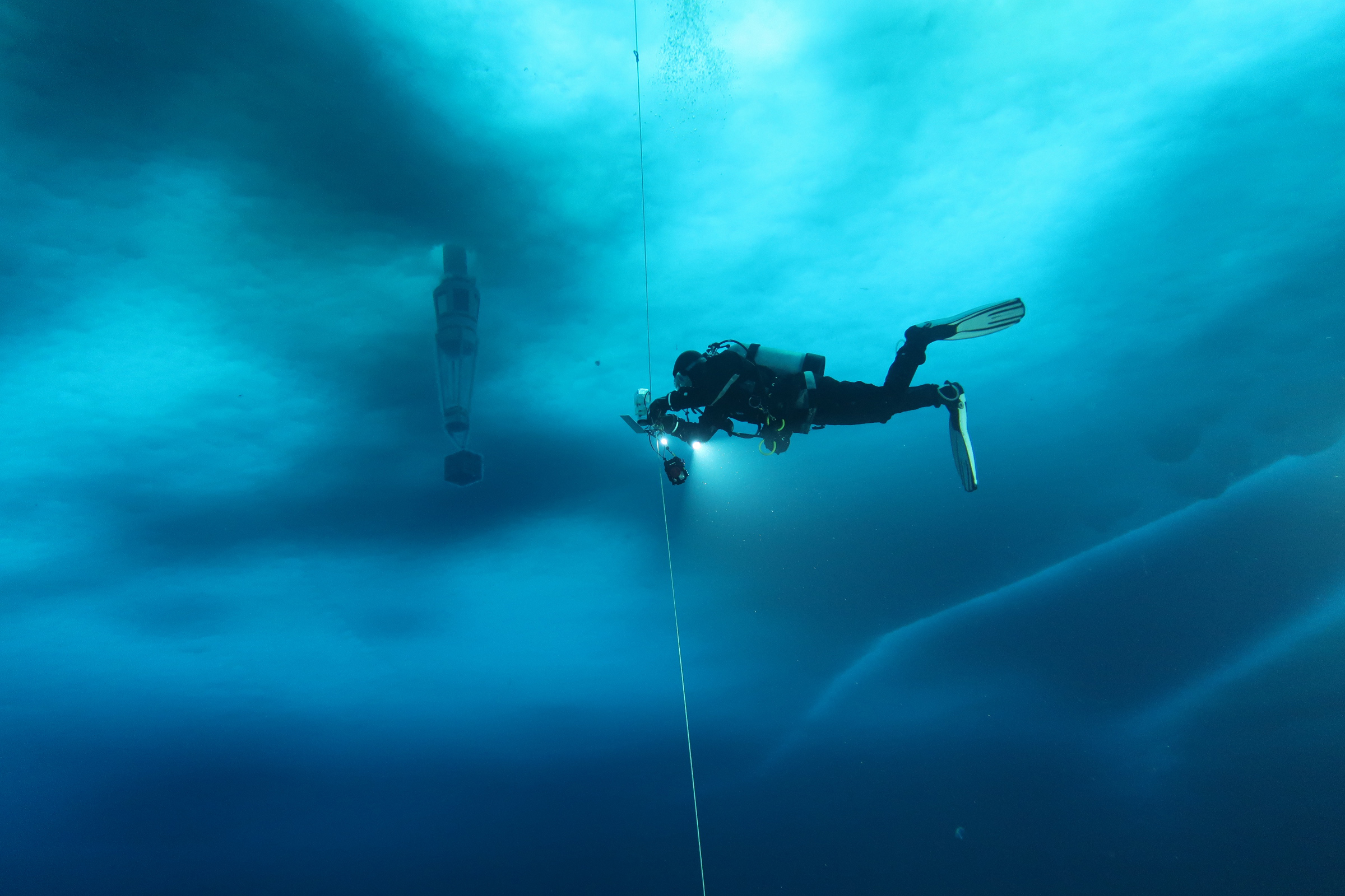 A diver under the ice swims towards an object suspended from the ice.