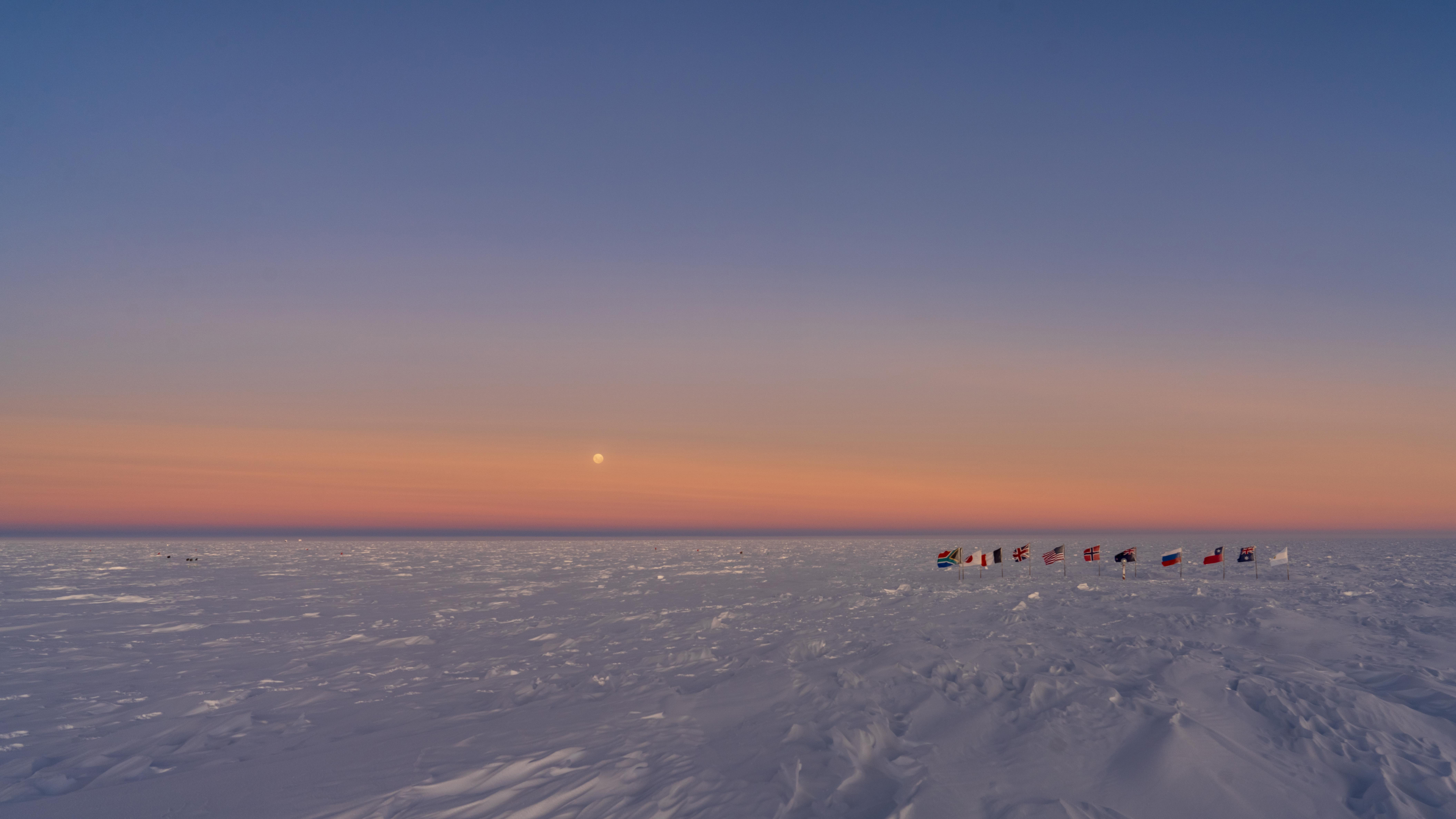 The moon shining through the sunrise-orange sky at the South Pole. At right are 12 national flags on short poles surrounding a candy can striped pole