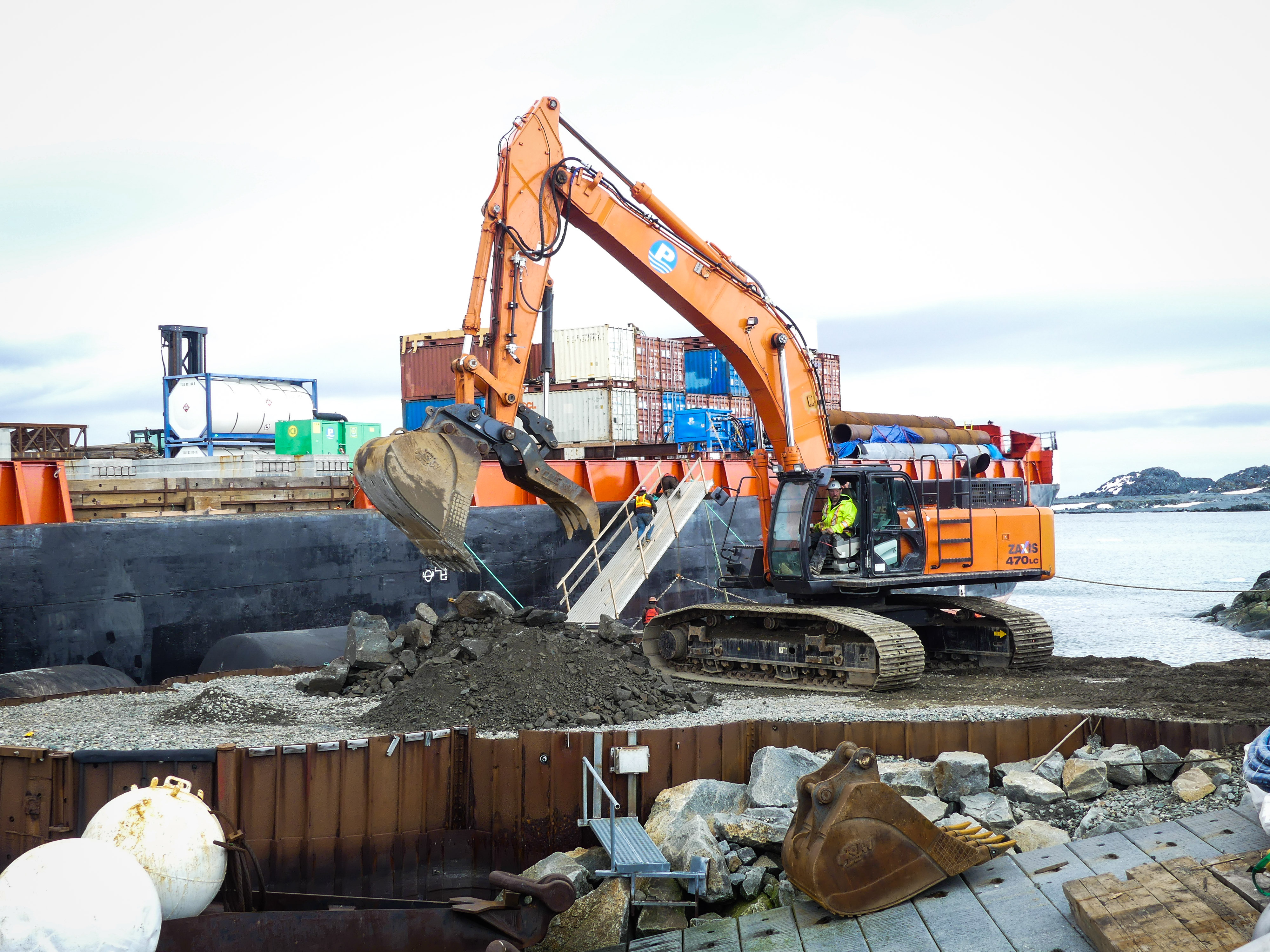 A backhoe excavates a dirt pier with a barge in the background. 