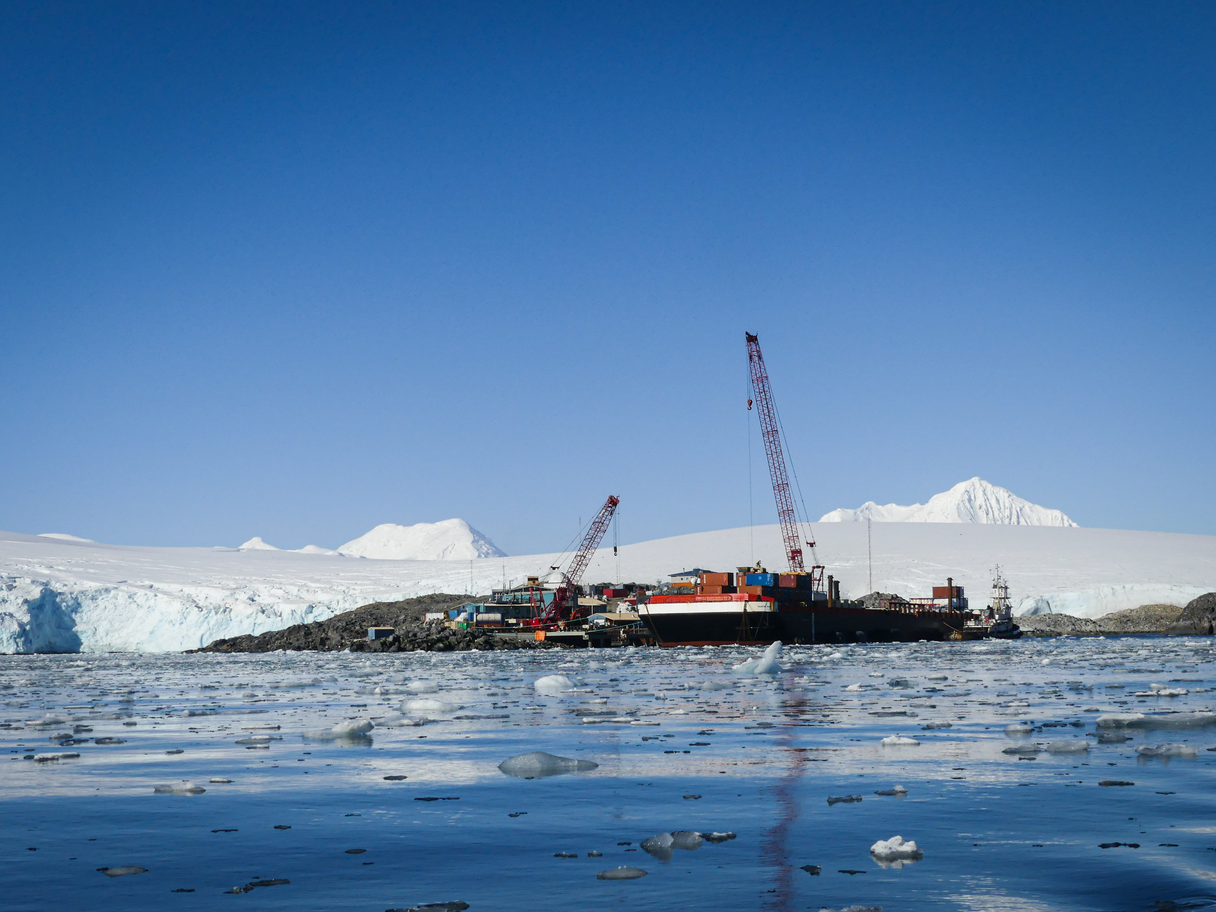 Construction cranes, a barge, and buildings with icy water in the foreground and mountainous glaciers behind them. 