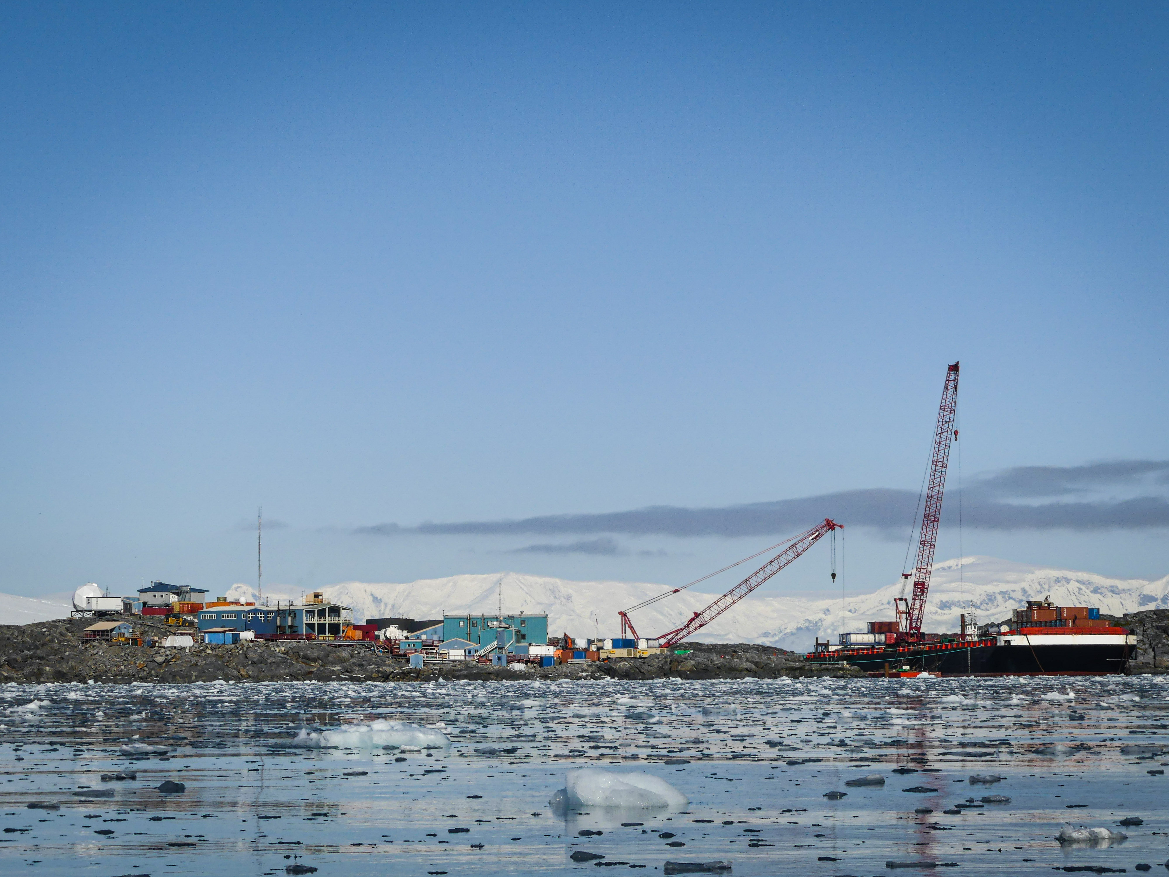 Construction cranes, a barge, and buildings with icy water in the foreground and mountainous glaciers behind them. 