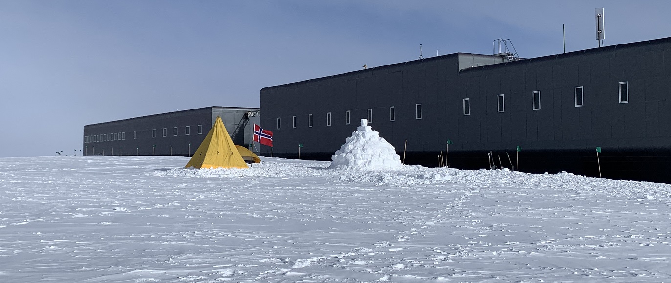 A yellow tent sits in front of a long gray building on snow.