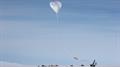 A scientific balloon rises above crane and other vehicles against light blue sky